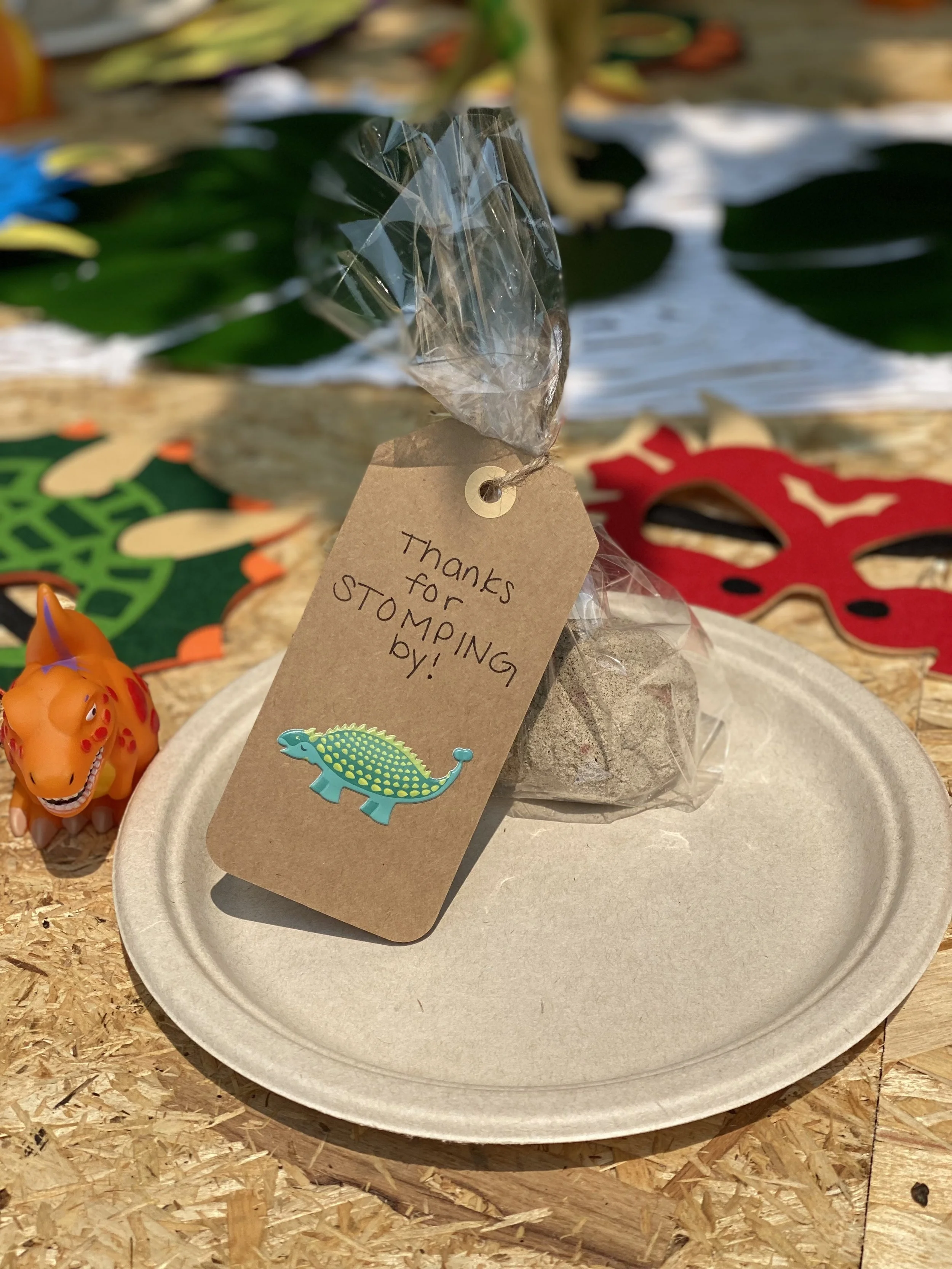 A small paper plate with a clear plastic bag containing a truffle or chocolate, tied with a string that has a brown tag saying 'Thanks for stomping by!' with a dinosaur sticker. There are colorful party masks and a toy dinosaur beside the plate, on a
