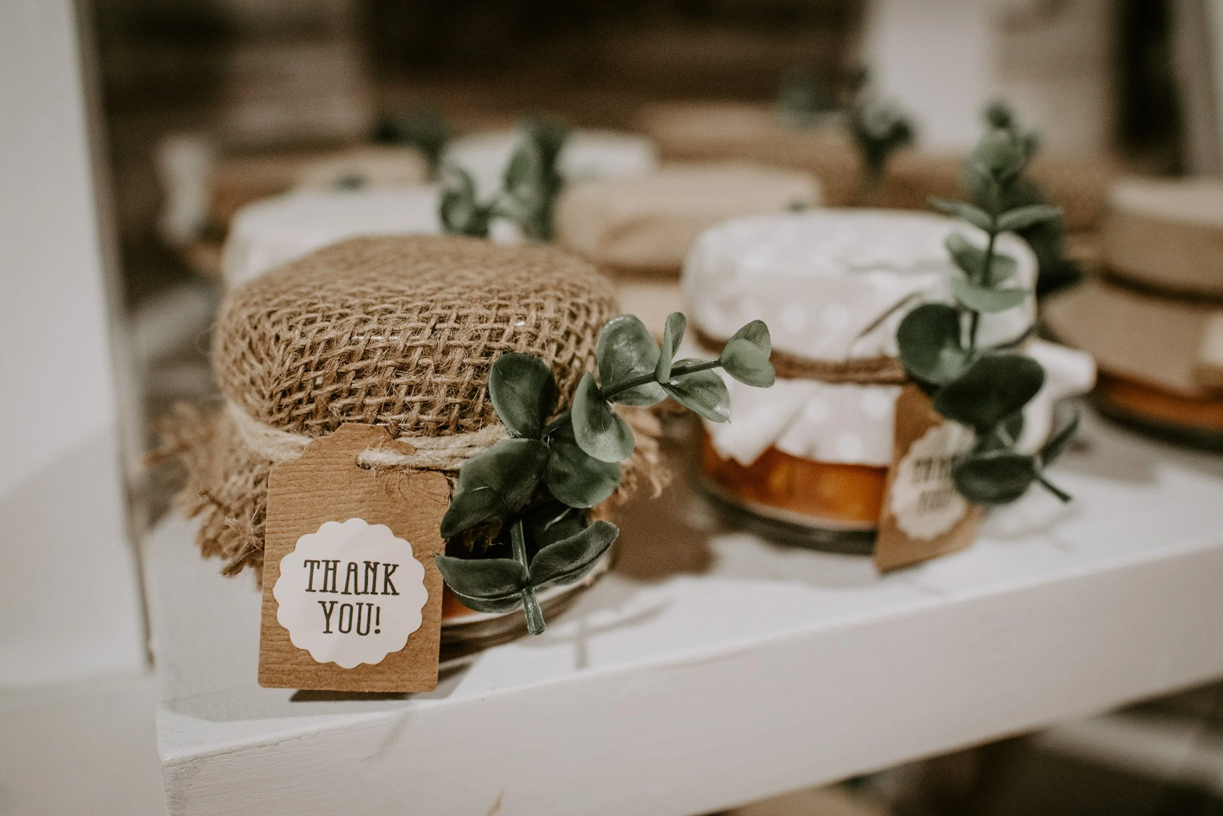 Three small jars with fabric and burlap covers, decorated with greenery, on a white shelf. One jar has a Thank You! tag attached.