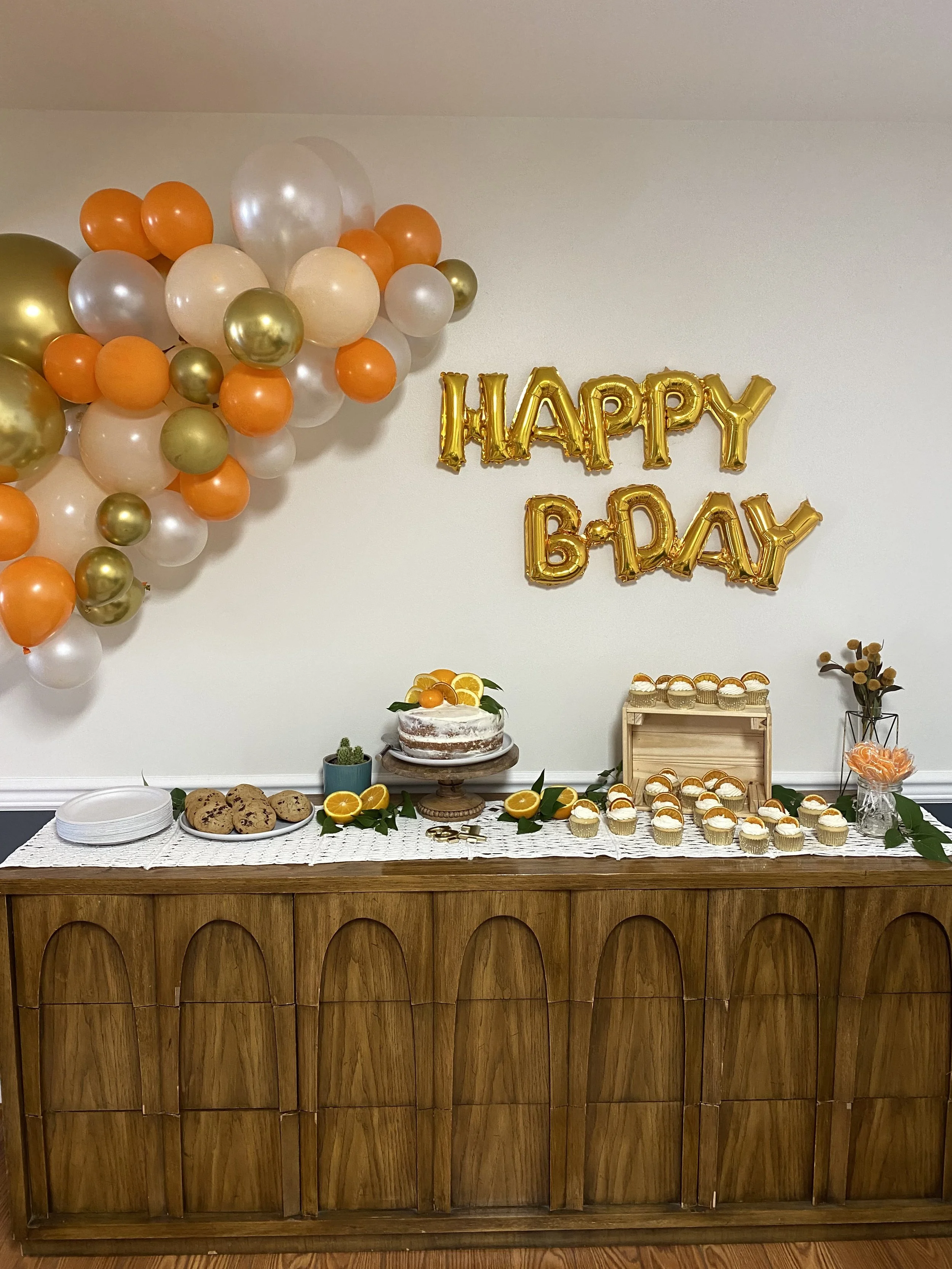 A birthday party setup with a balloon decoration wall featuring orange, white, gold, and metallic balloons. A wooden table is adorned with cookies, cupcakes, a cake, and orange slices, with a 'HAPPY B-DAY' gold balloon banner on the wall behind.