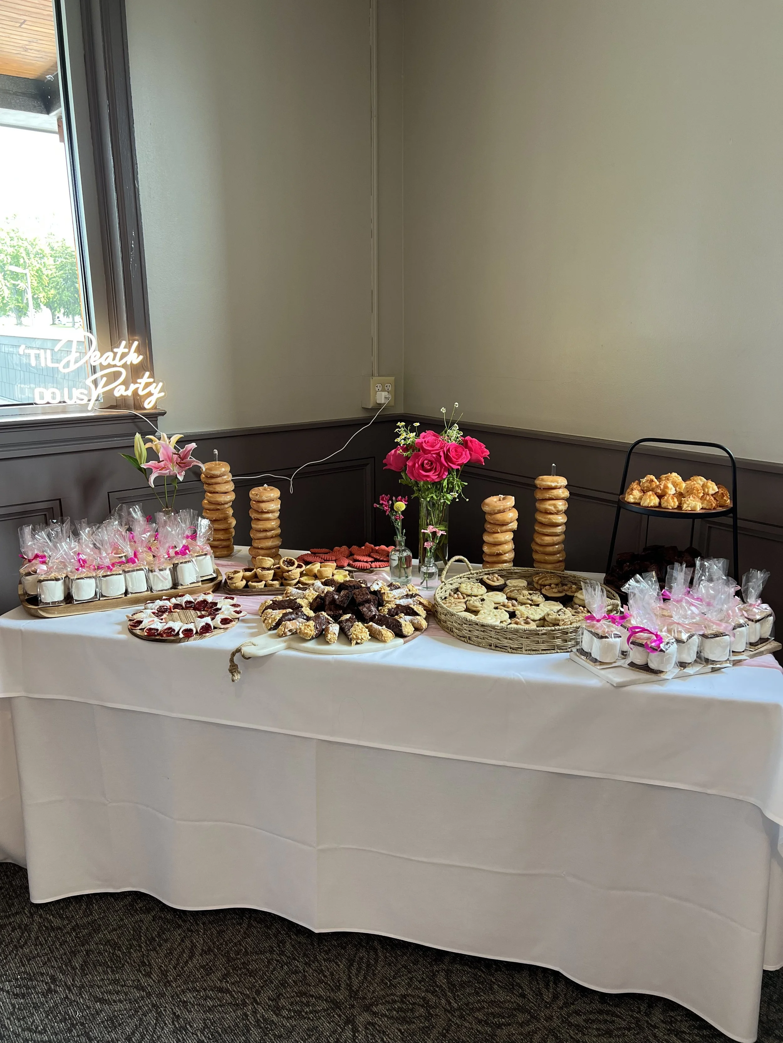 A dessert table with cookies, cupcakes, and candy, decorated with pink flowers and stacked donuts, in a room with beige walls and a window.