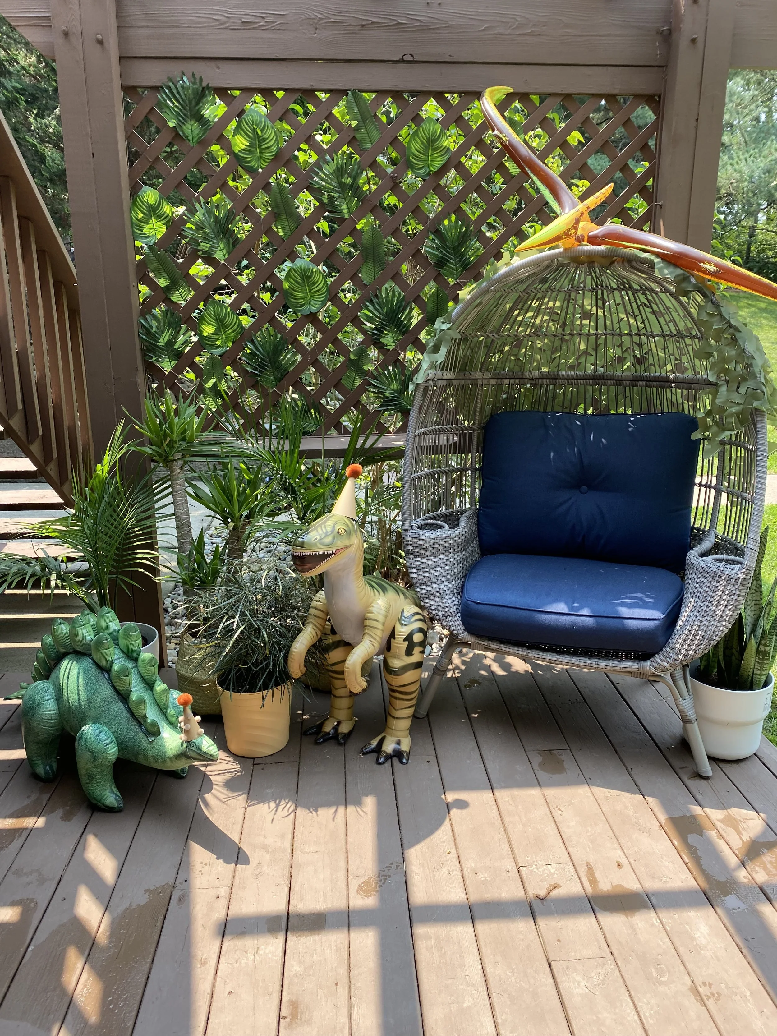 Decorative outdoor patio corner with artificial dinosaurs, potted plants, wicker chair with blue cushions, and a wooden lattice with green leaf decorations.