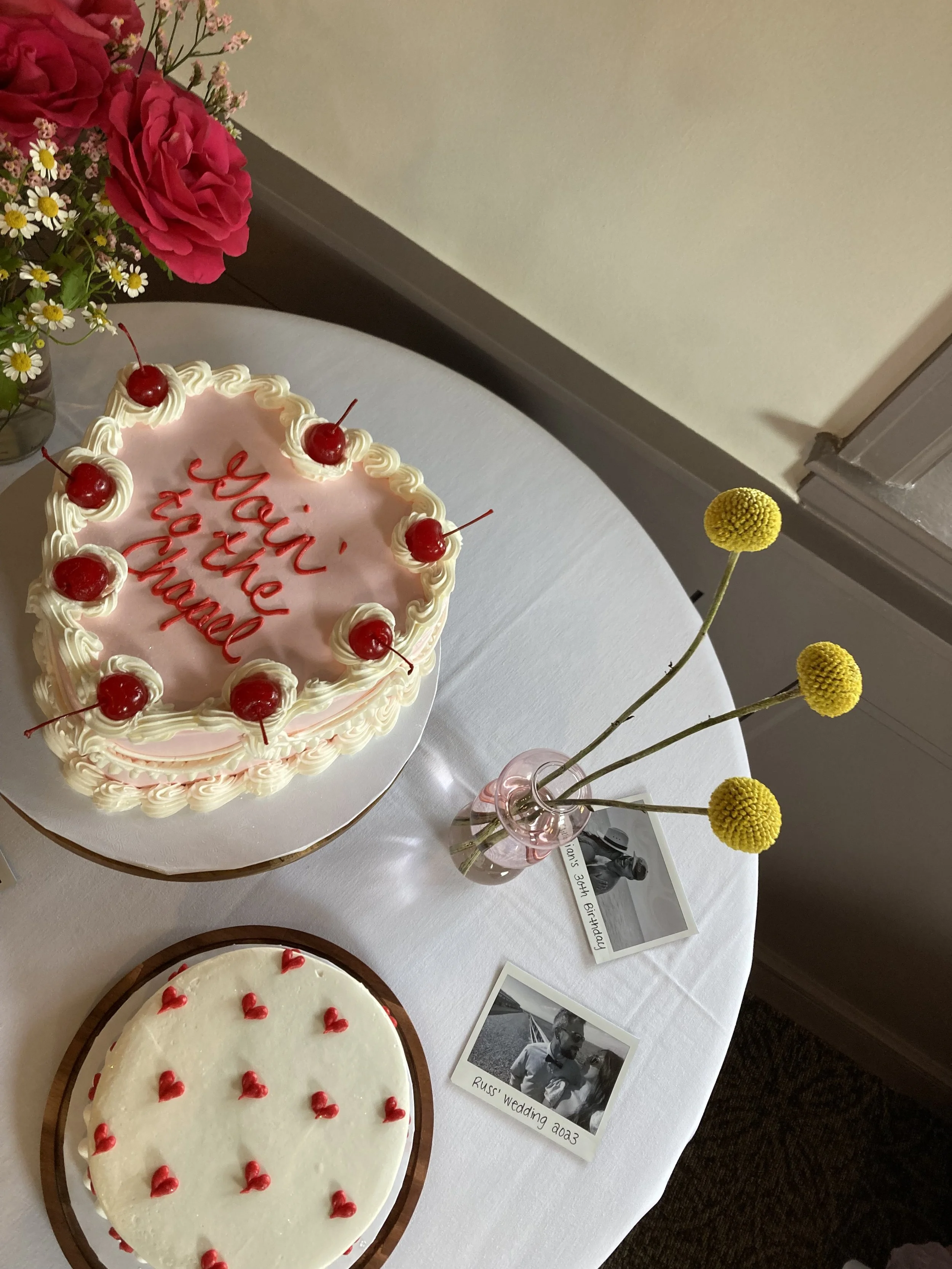 Two cakes are on a table, one with pink icing, cherries, and the words 'You're the best' written in red, and the other with white icing and small red hearts. A small vase with three yellow billy ball flowers is next to the cakes, and there are two bl