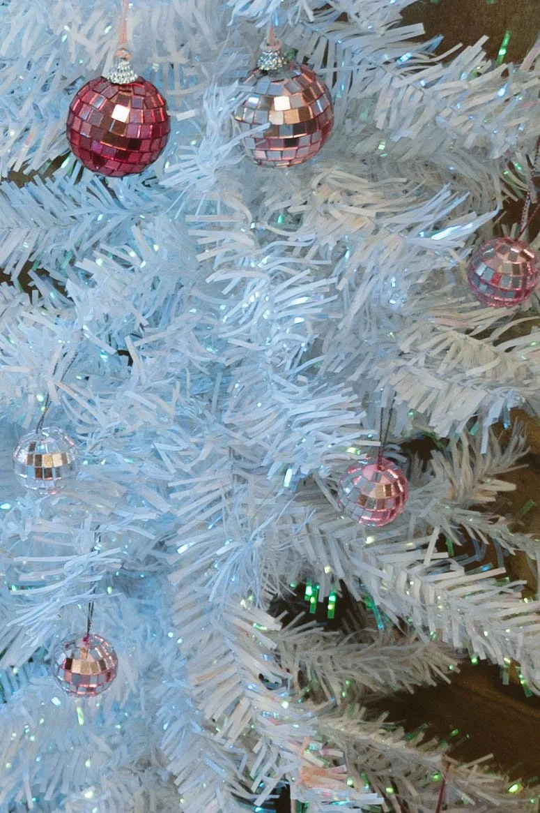 A white artificial Christmas tree decorated with small pink and silver disco ball ornaments and string lights.