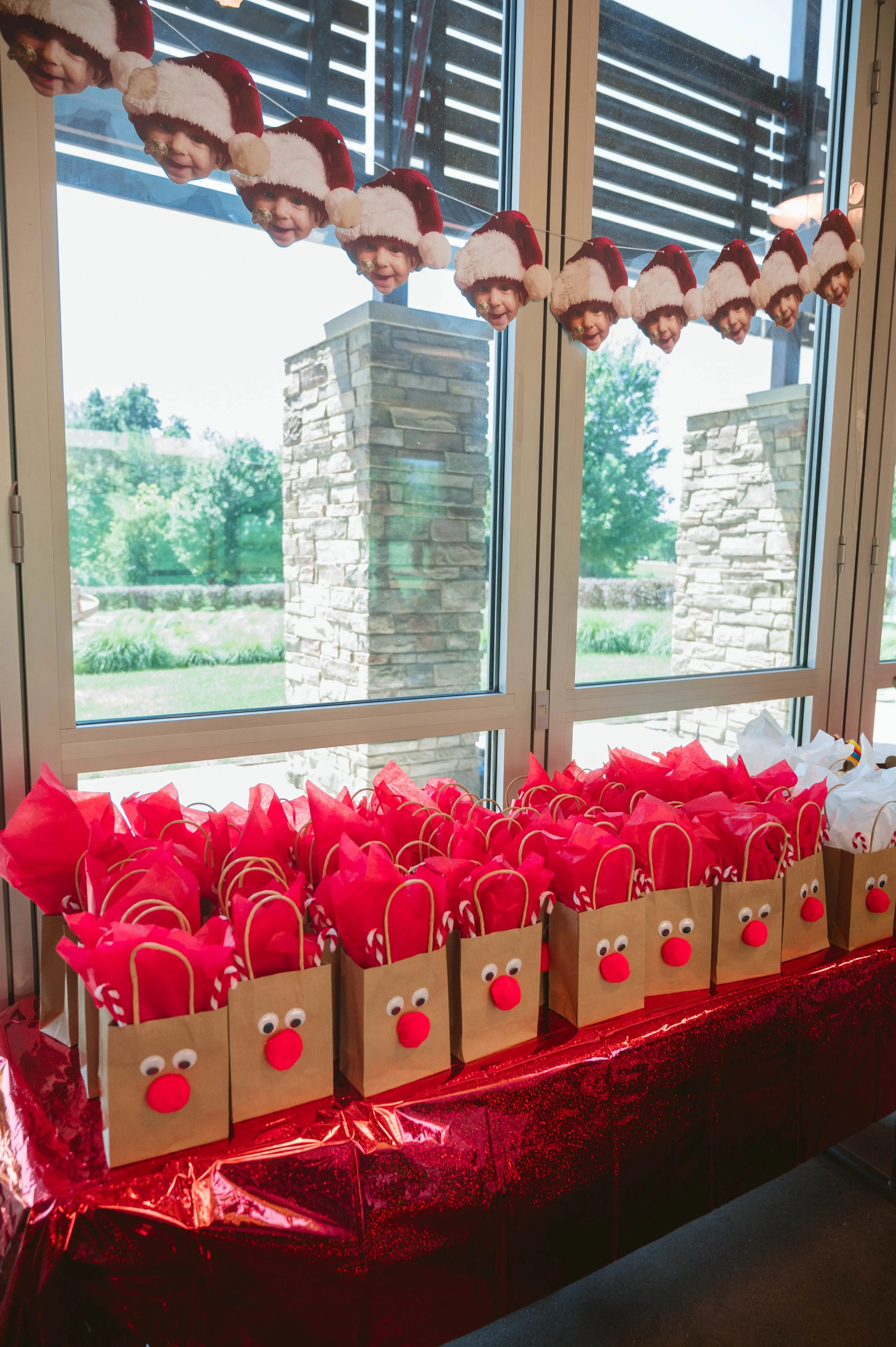 Holiday party table with gift bags decorated with reindeer faces, felt red noses, googly eyes, and candy cane handles, placed on a red tablecloth in front of a window with Santa hat garland hanging above.