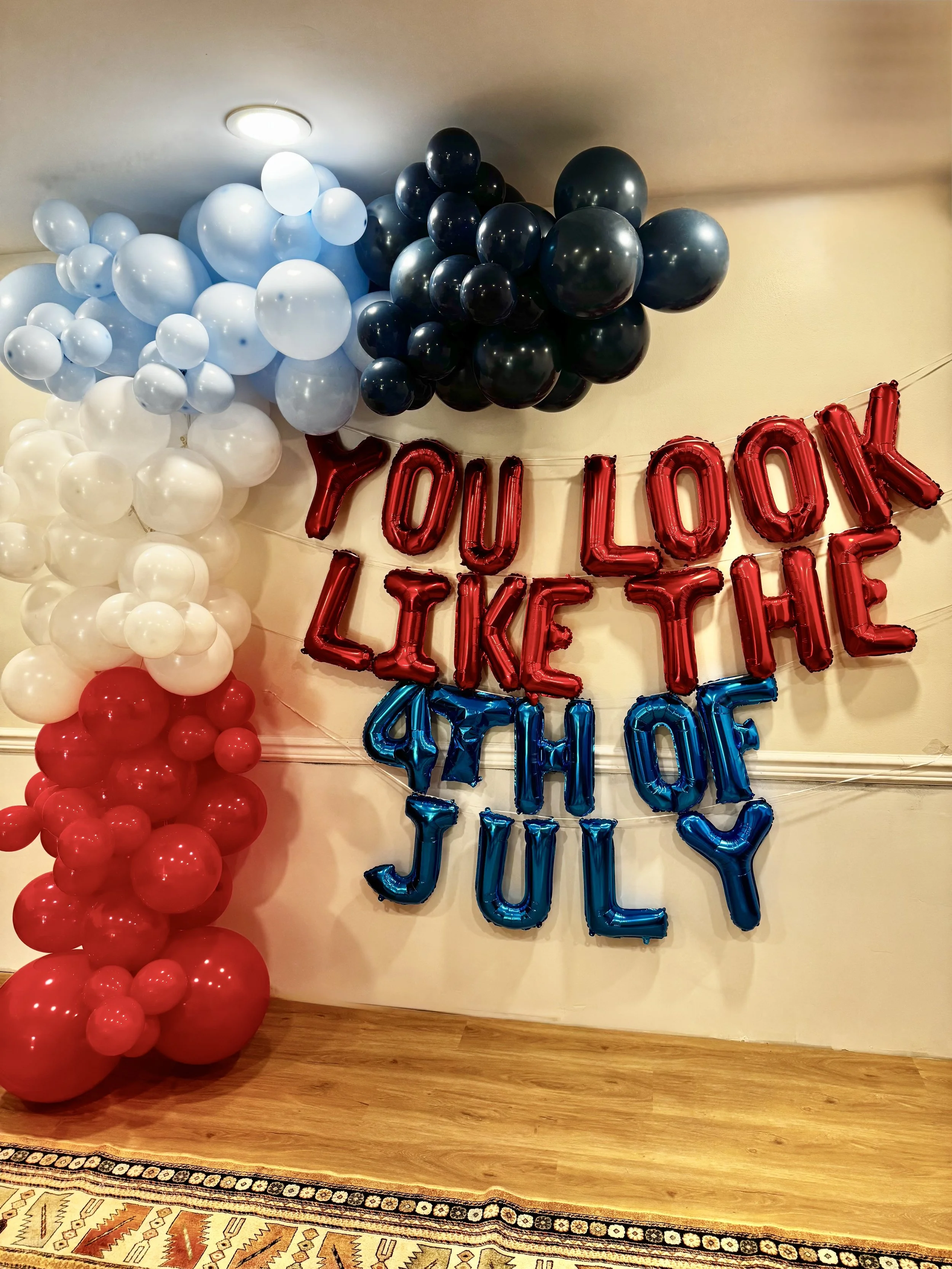 Decorative balloon arrangement with red, white, and black balloons on a wall, featuring a red and blue balloon banner that reads 'You Look Like The 4th Of July'.