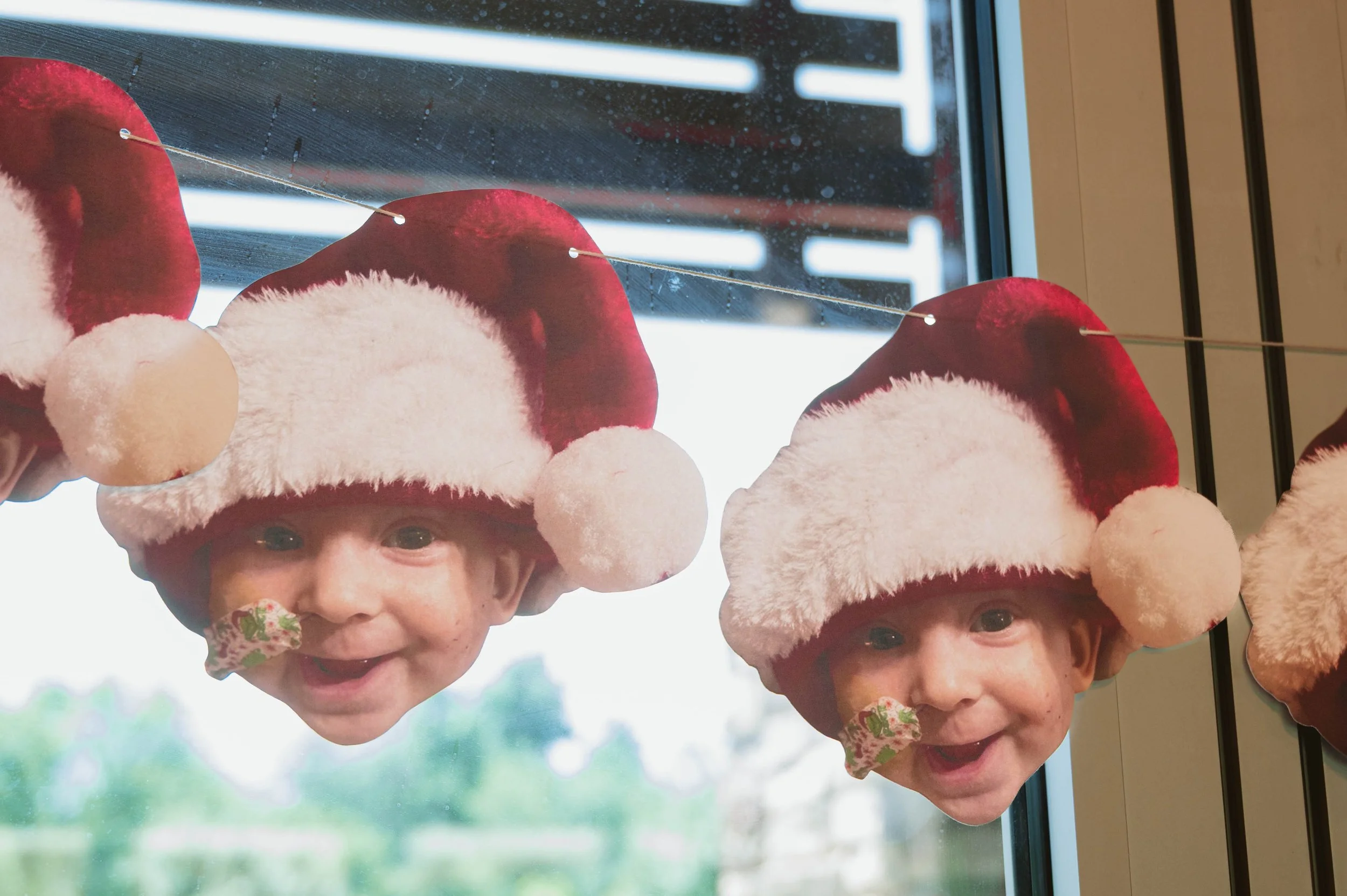 Decorative Christmas ornaments featuring children's faces wearing Santa hats hanging on a string in front of a window.