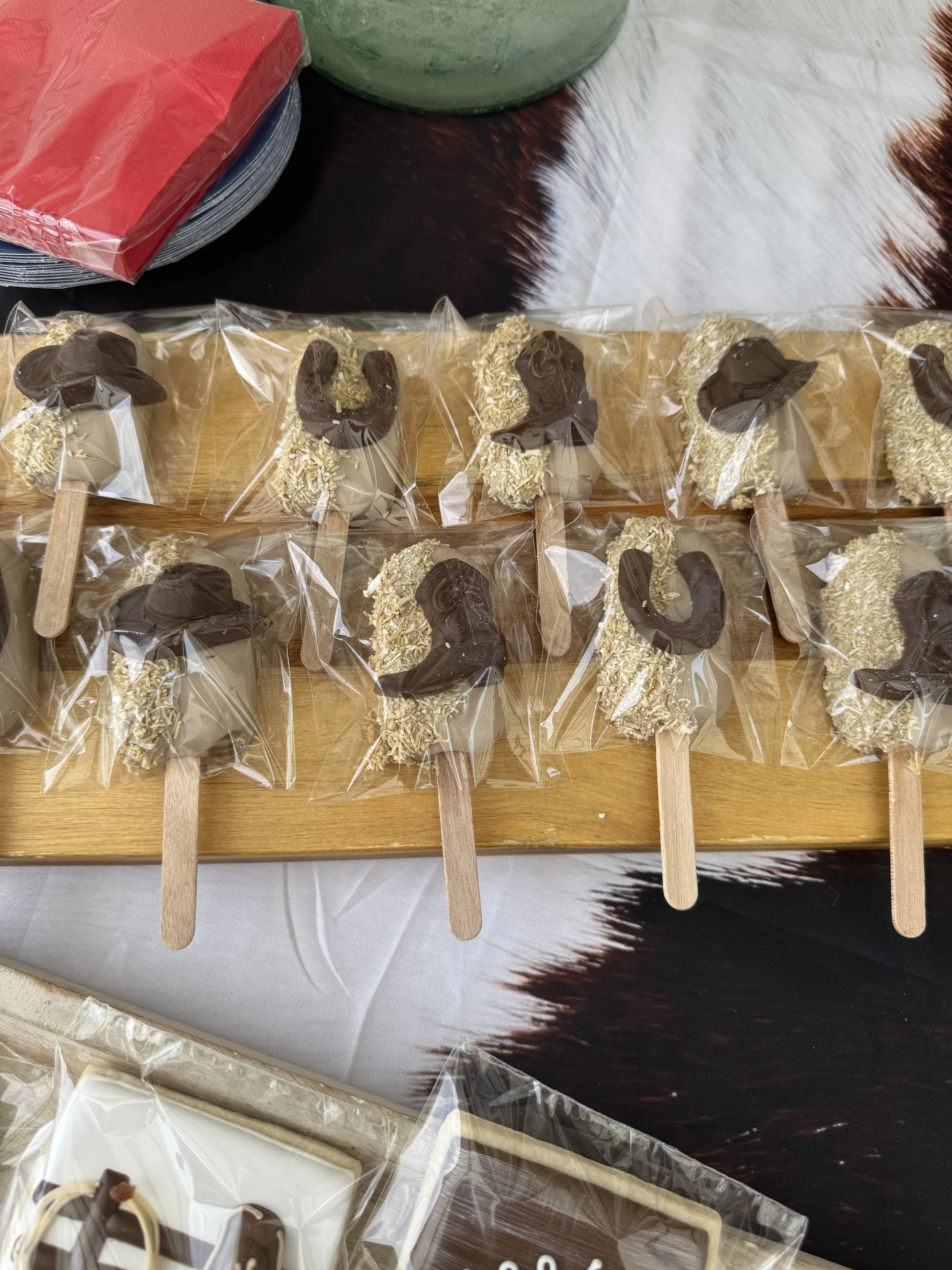 Ten painted dessert popsicles wrapped in clear plastic on a wooden tray, with chocolate and coconut toppings, and some red and green plates and a cowhide cloth in the background. baby shower, cowboy themed bay shower, cakesciles, cowboy themed cookie