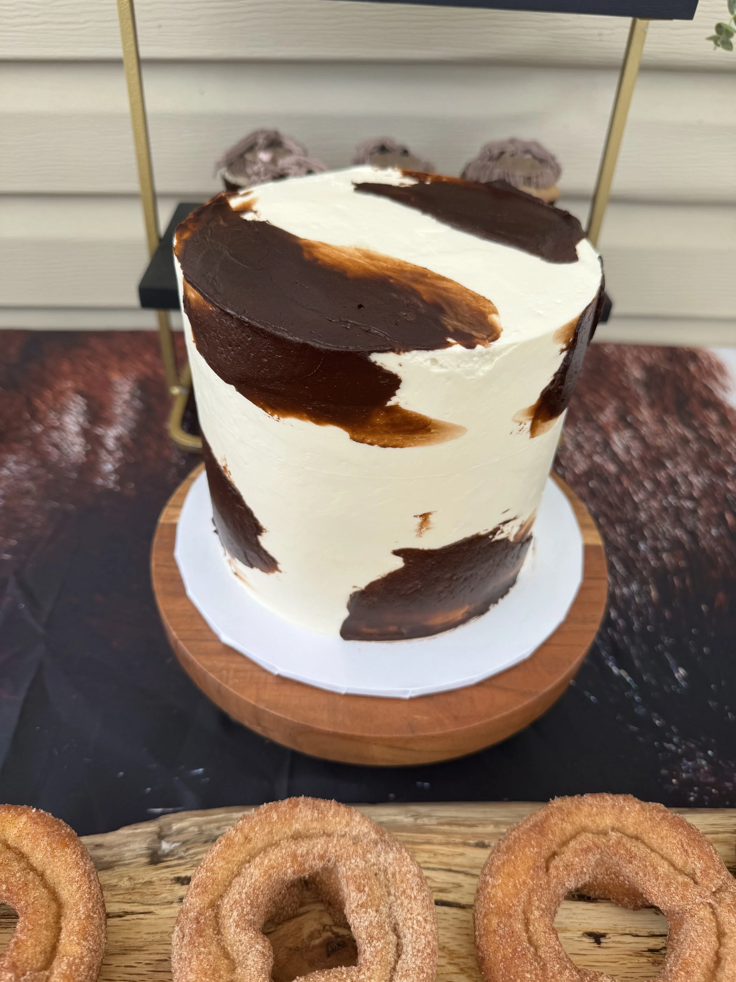 A tall cake with white frosting and large brown and black abstract chocolate design on top, placed on a round wooden cake stand. In front, there are three golden-brown cinnamon sugar donuts on a wooden tray.