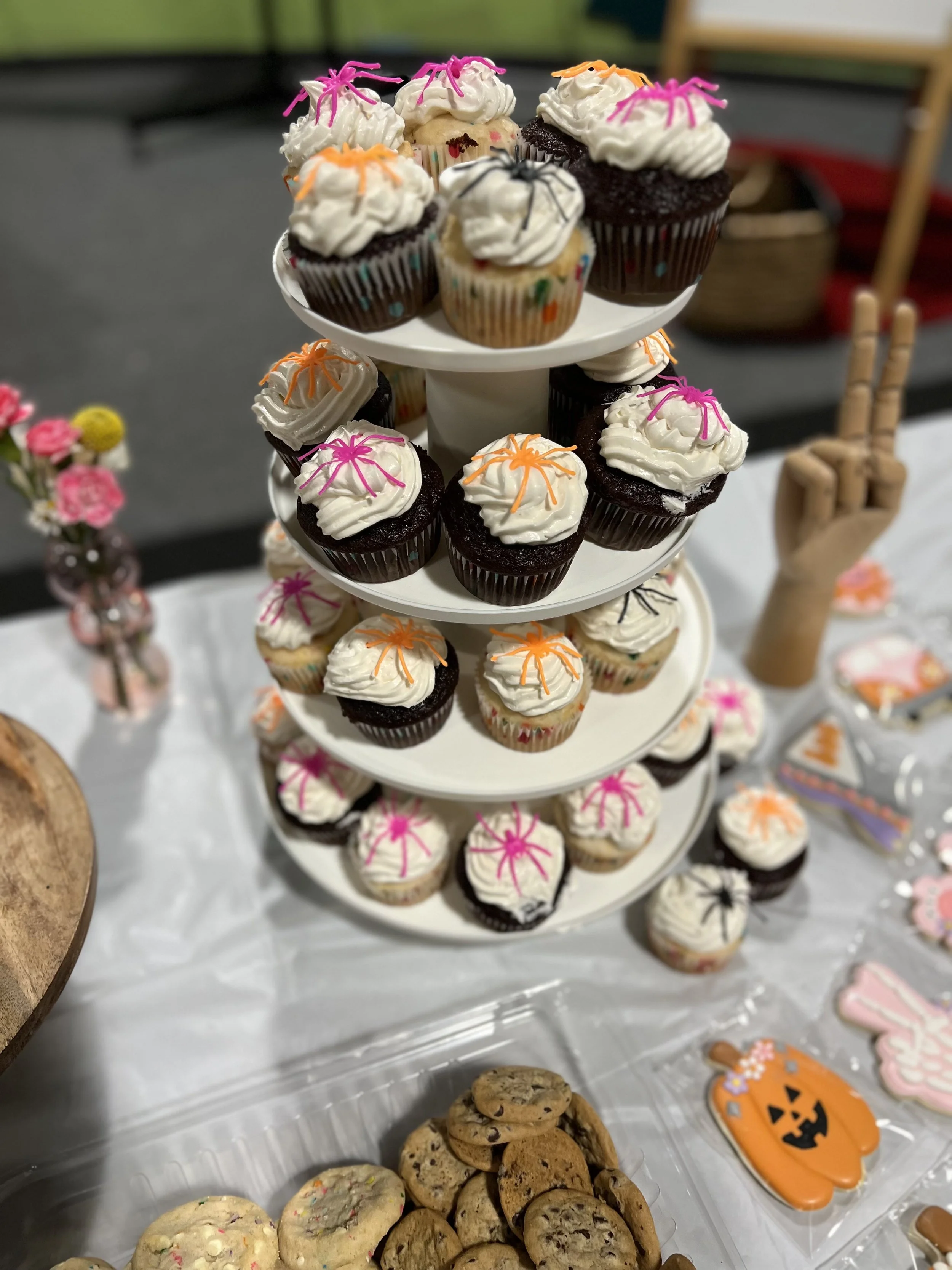 A three-tiered dessert stand filled with decorated cupcakes. The cupcakes have white frosting and colorful decorations such as pink, orange, and black spider-like sprinkles. There are additional cookies and Halloween-themed decorations on the table.