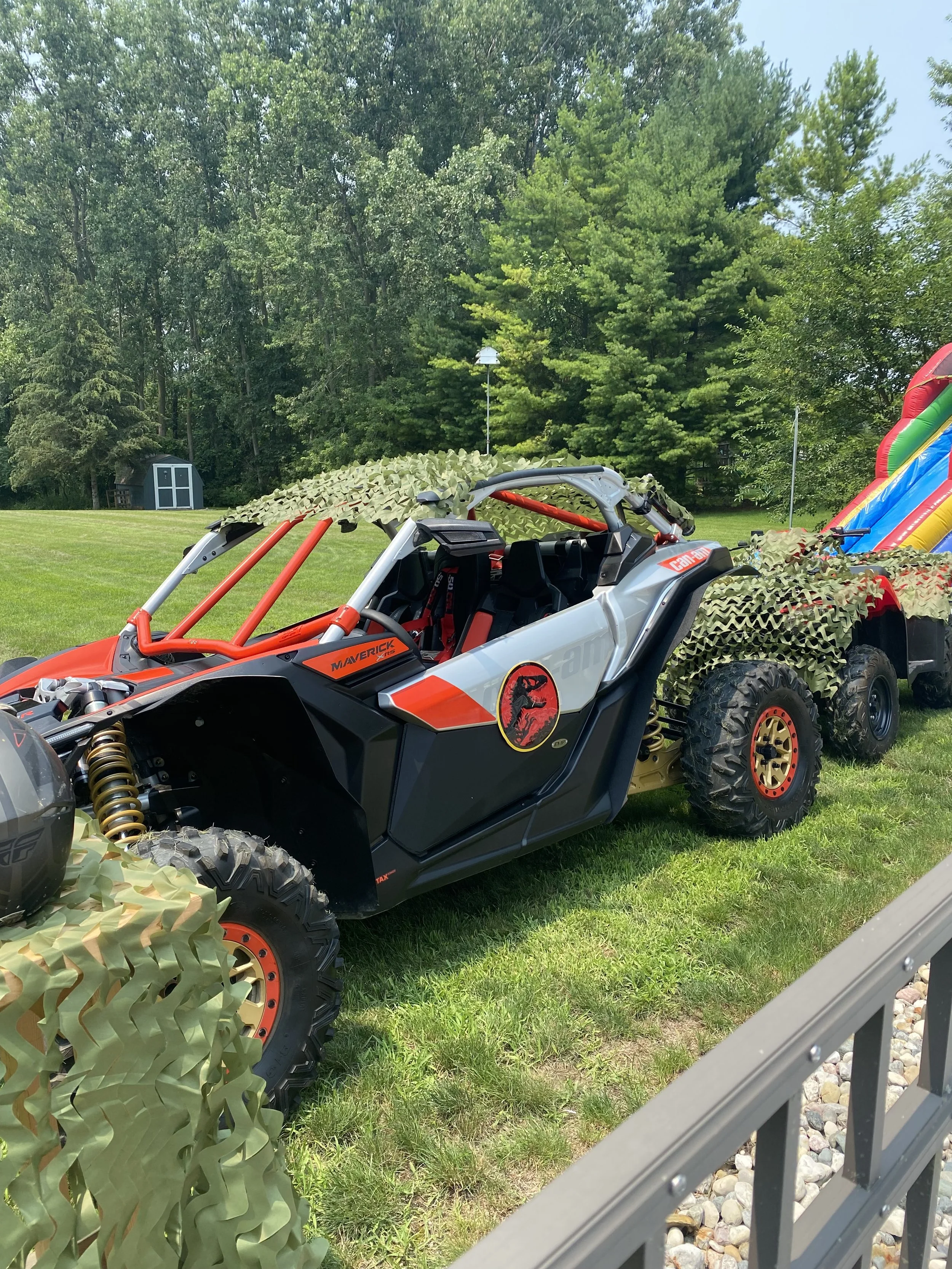 A toy car designed to resemble a dinosaur, with emerald green camouflage netting on top and behind, is parked on a grassy yard. The car features a Jurassic Park logo and a red and black color scheme.