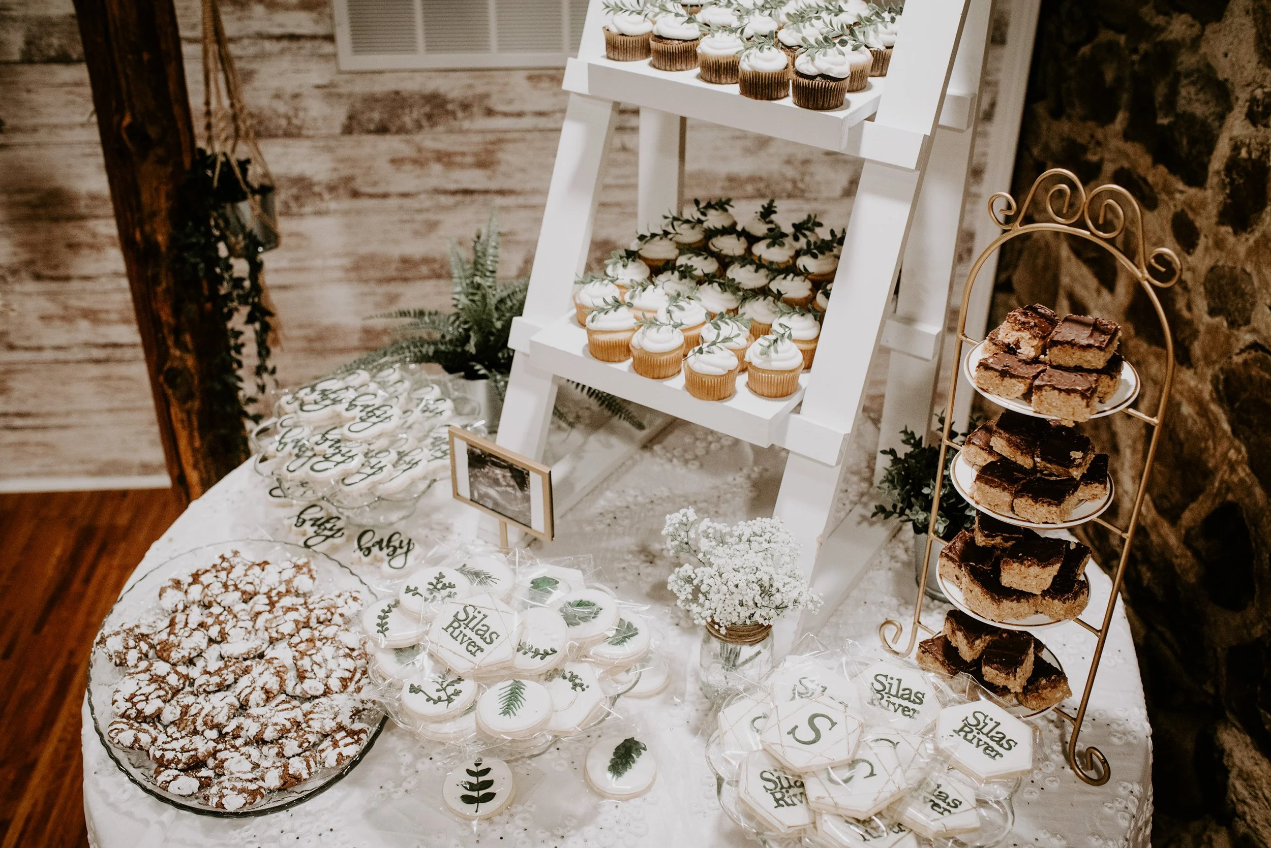 Display of various cookies and cupcakes at a dessert table, with some labeled cookies that read 'Silas River,' and decorated with greenery for a celebration.