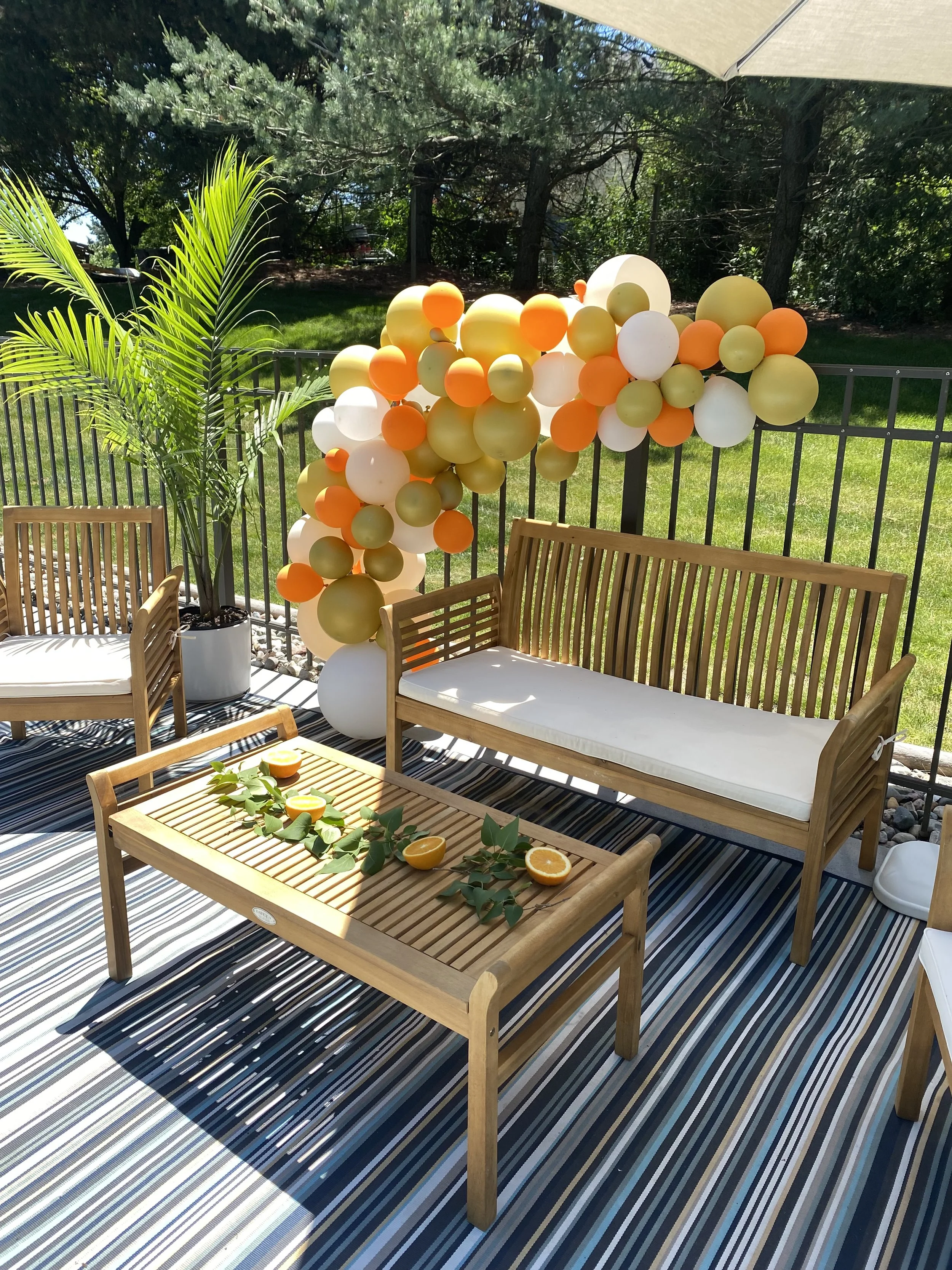 Outdoor patio decorated with a balloon arch in shades of yellow, white, orange, and green, with wooden furniture including a bench and chairs, a striped rug, a potted palm plant, and a table with orange slices and green leaves.