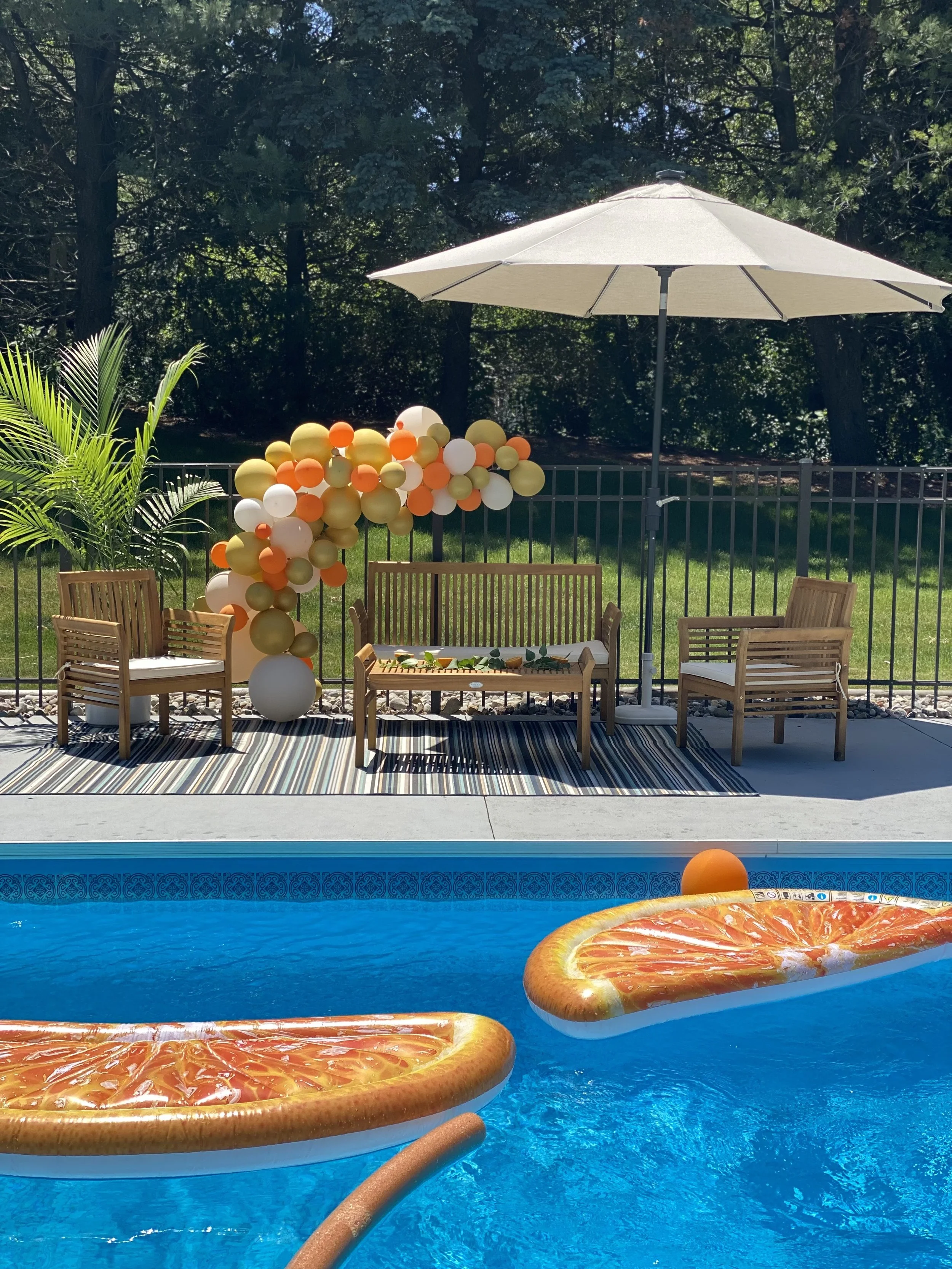 Poolside area with outdoor furniture, a large white umbrella, a balloon decoration arch, and an inflatable orange slice float in the pool.