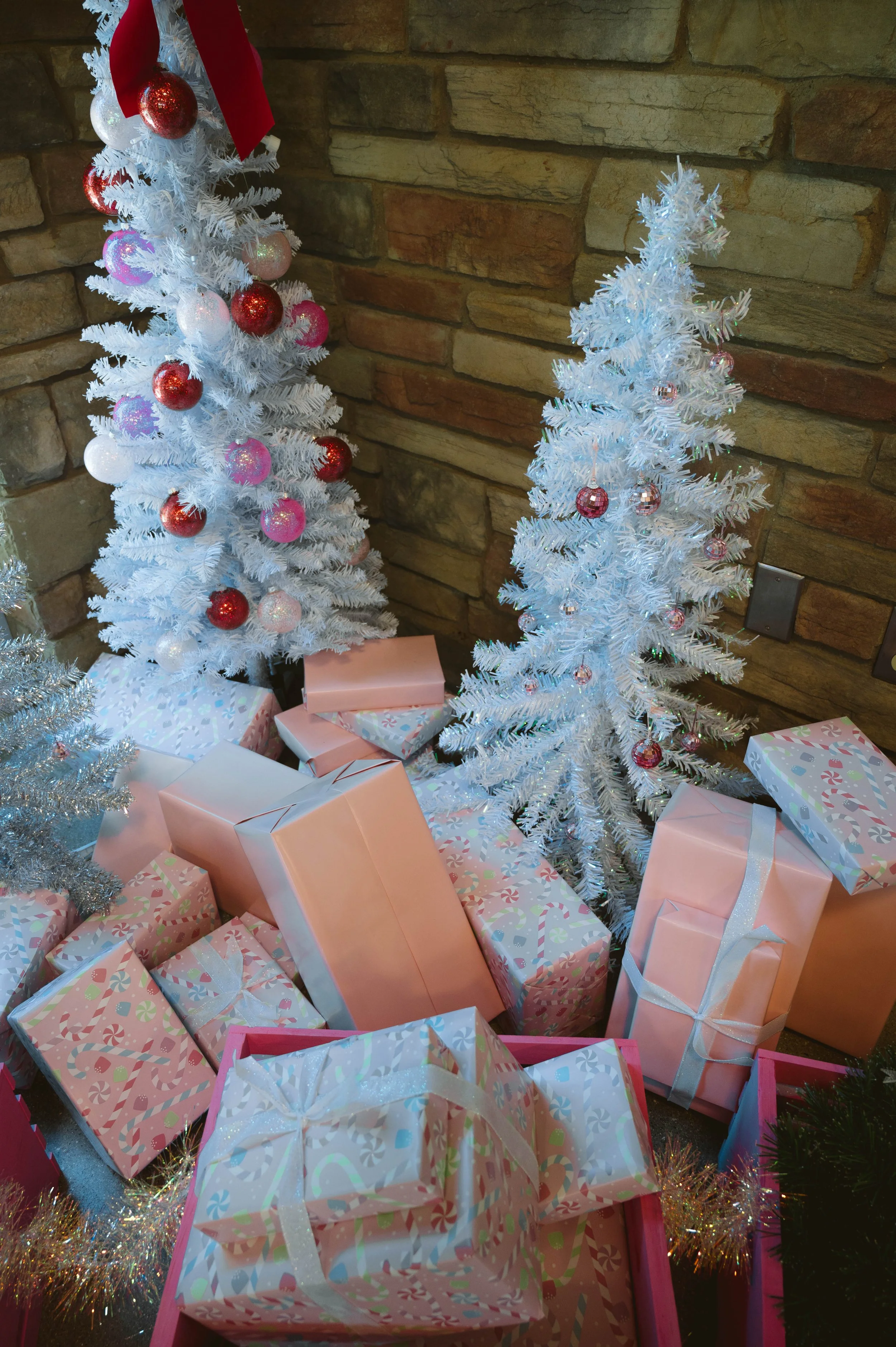 Decorative Christmas scene with two white artificial Christmas trees, one adorned with red, pink, and white ornaments, surrounded by wrapped gifts in pastel and patterned wrapping paper on a wood-paneled wall background.