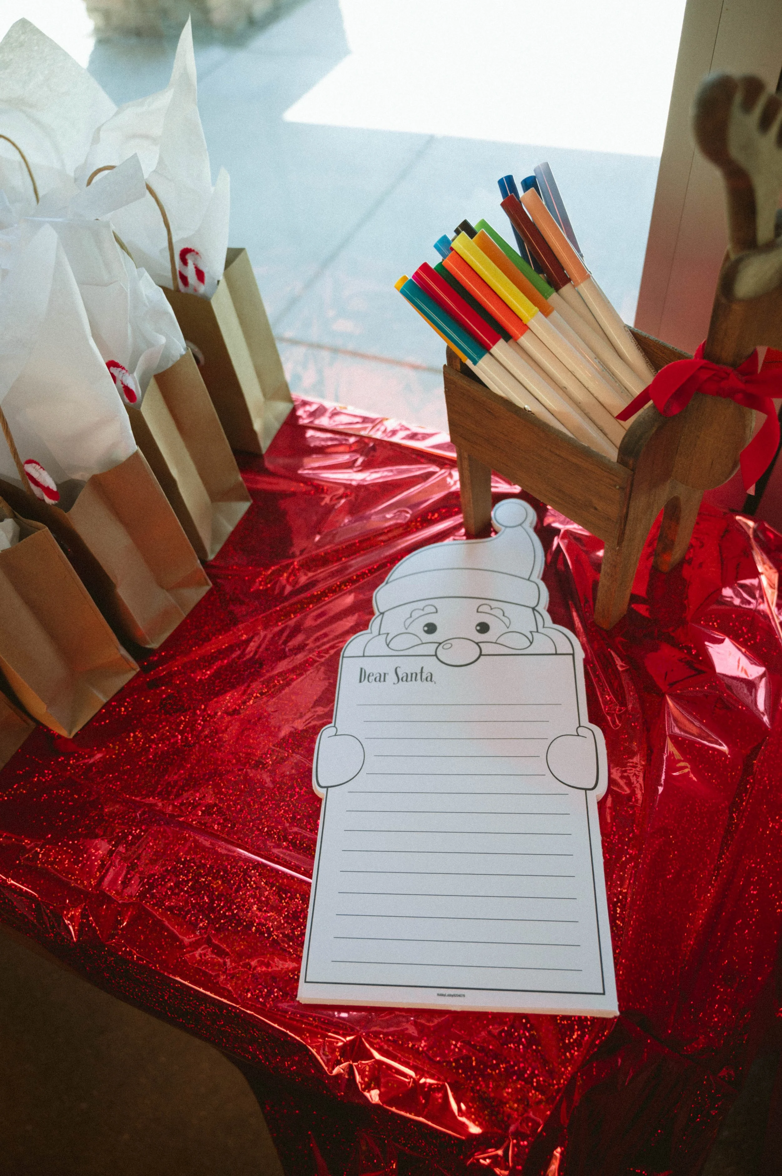 A Christmas-themed table with a red, sparkly tablecloth, holding a Santa letter template, a small wooden reindeer, a wooden container filled with colorful pens, and paper bags with red and white holiday decorations.