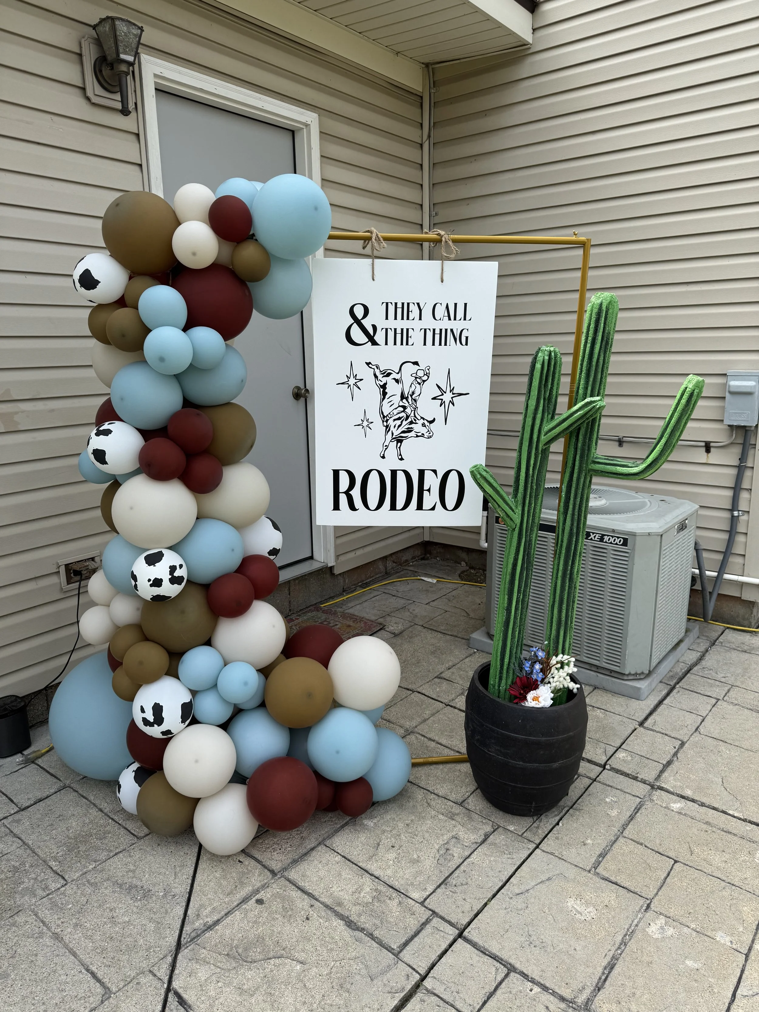 Decorations for a rodeo event featuring balloons in cow print, white, light blue, maroon, and brown, a sign with a cow graphic and the text '& They Call the Thing Rodeo', and a large faux cactus in a black pot with flowers, set on a paved patio next