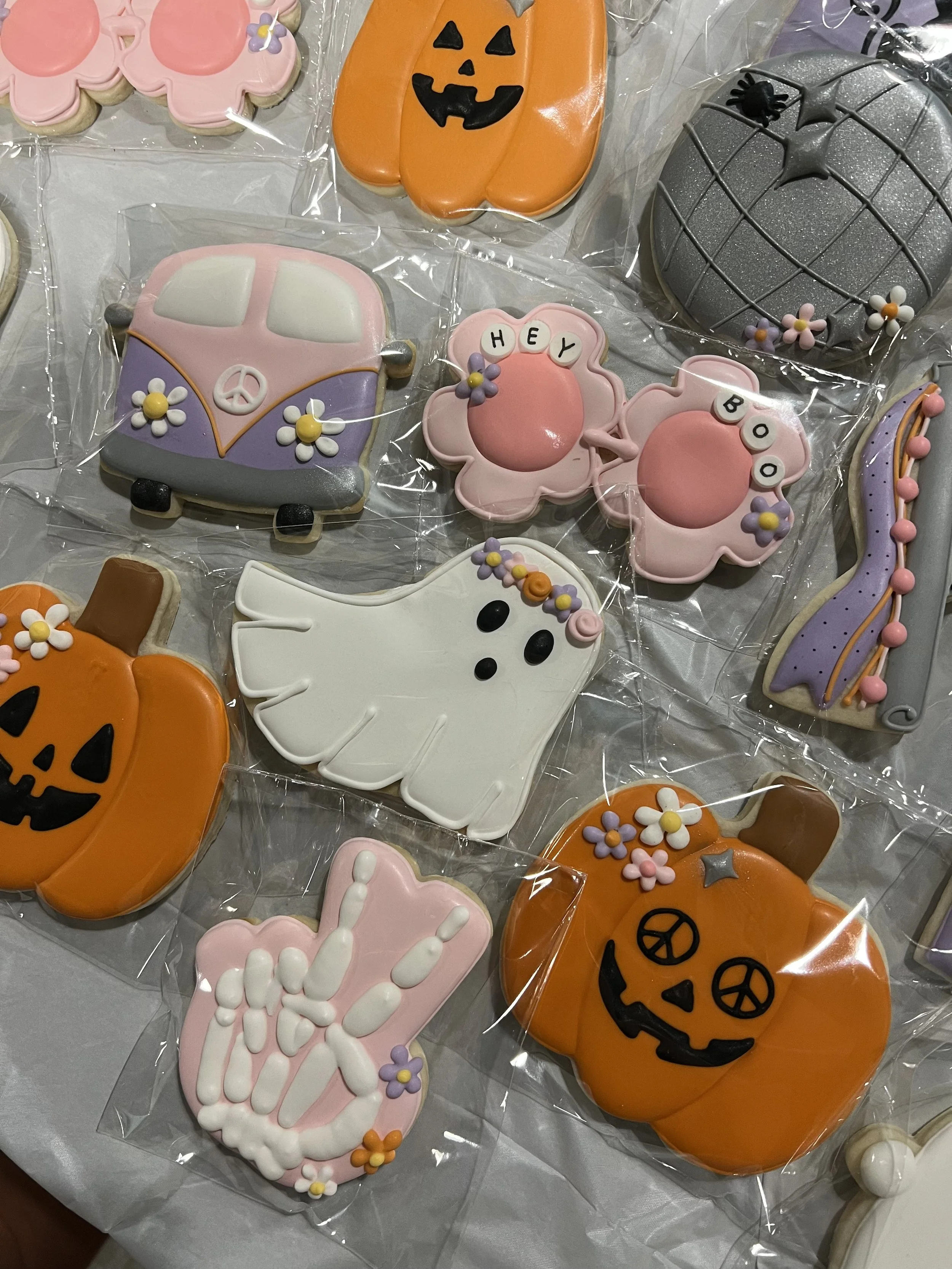Assorted Halloween-themed decorated cookies, including pumpkins, a ghost, a peace sign hand, a van, and phrases like 'HEY' and 'BOO', decorated with colorful icing and small sugar flowers.
