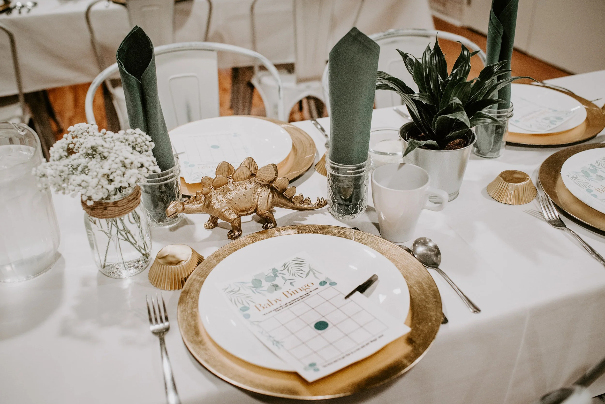 Table setting with gold-rimmed plates, green napkins, white cups, and decorative items including a small potted plant, plastic dinosaur figurine, and a baby bingo card on a white tablecloth.