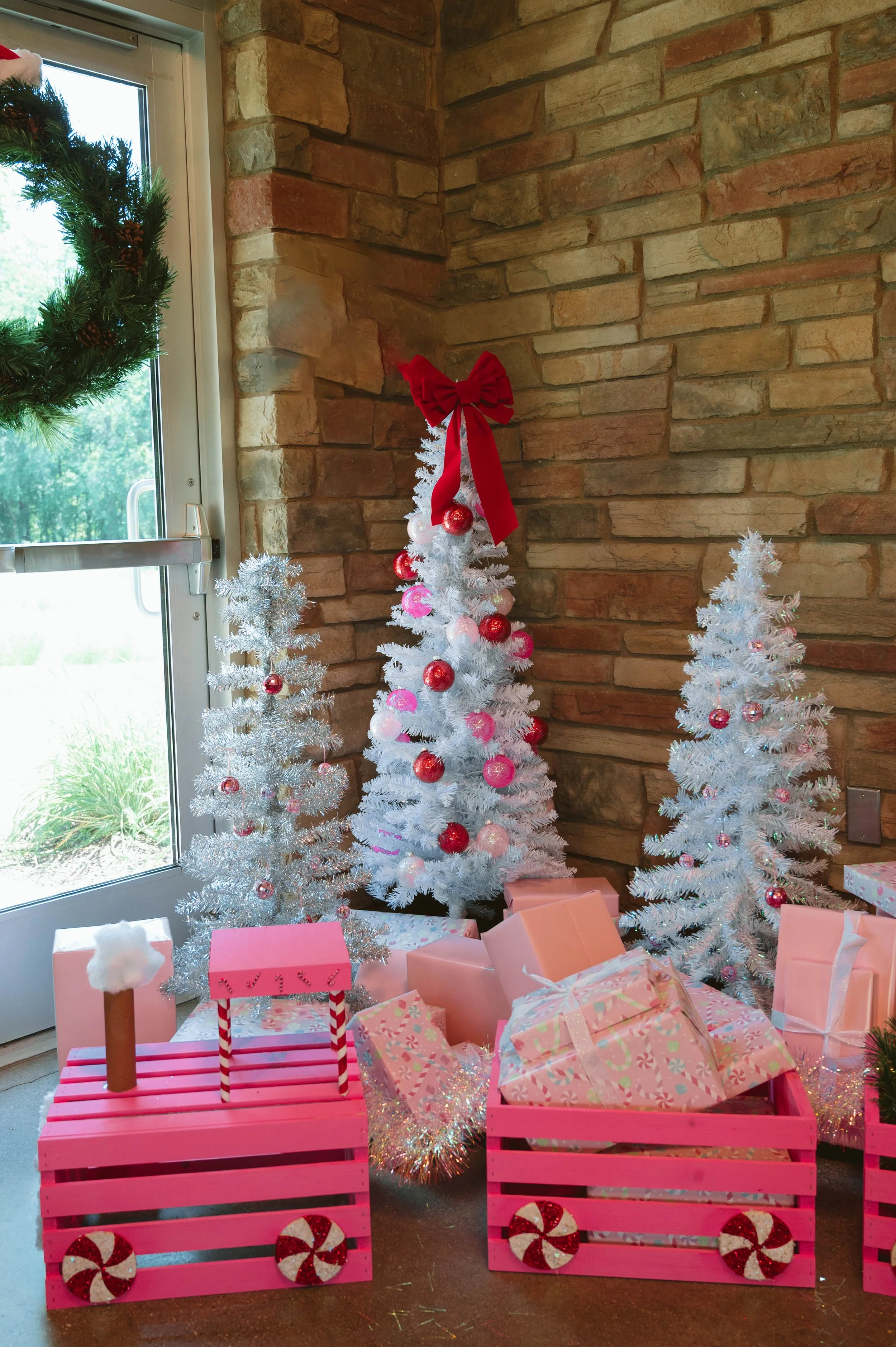 Decorative Christmas scene with three white and pink decorated Christmas trees, pink crates filled with wrapped presents and a candy cane decoration, set against a brick wall near a window.