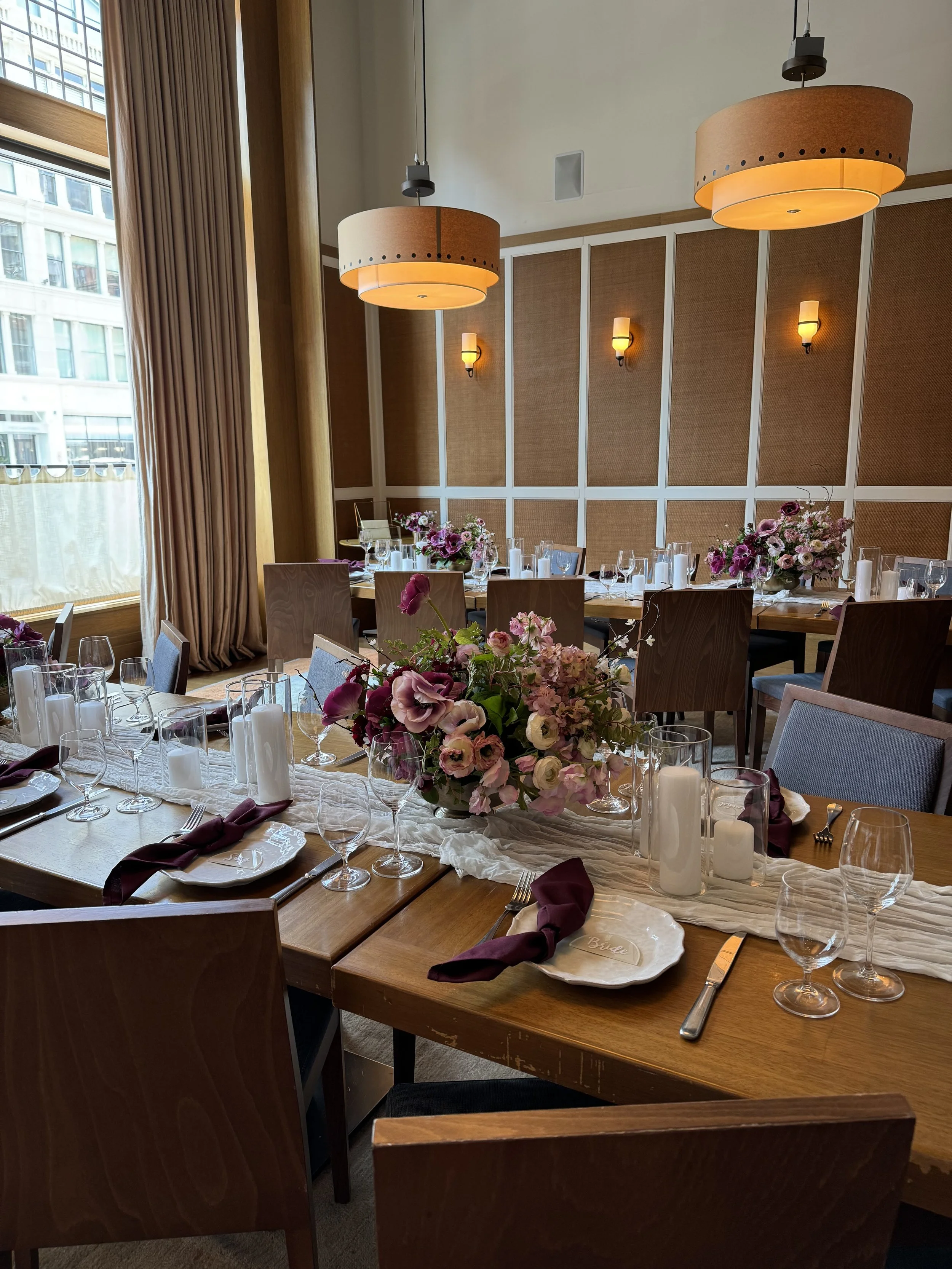 Elegant dining table decorated with a pink and purple floral centerpiece, candles, glassware, plates, and folded napkins in a brightly lit restaurant with large windows and hanging lamps.