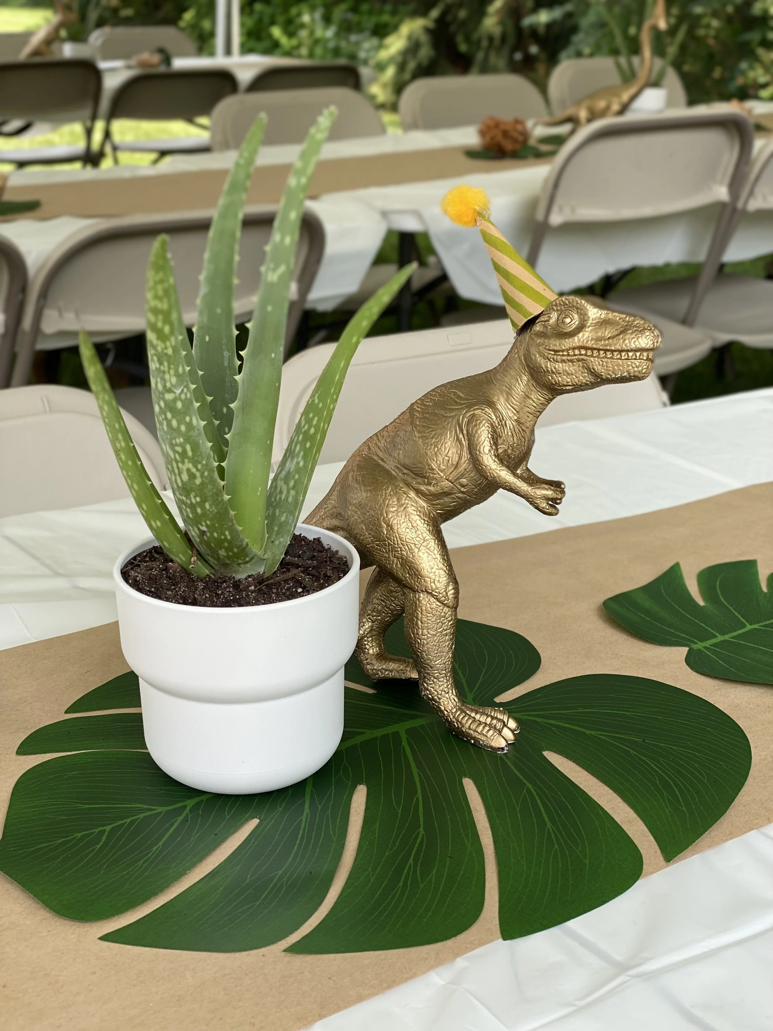 Gold-colored dinosaur figurine wearing a party hat, seated behind a potted aloe vera plant on a decorative green leaf, with tables and chairs in the background.