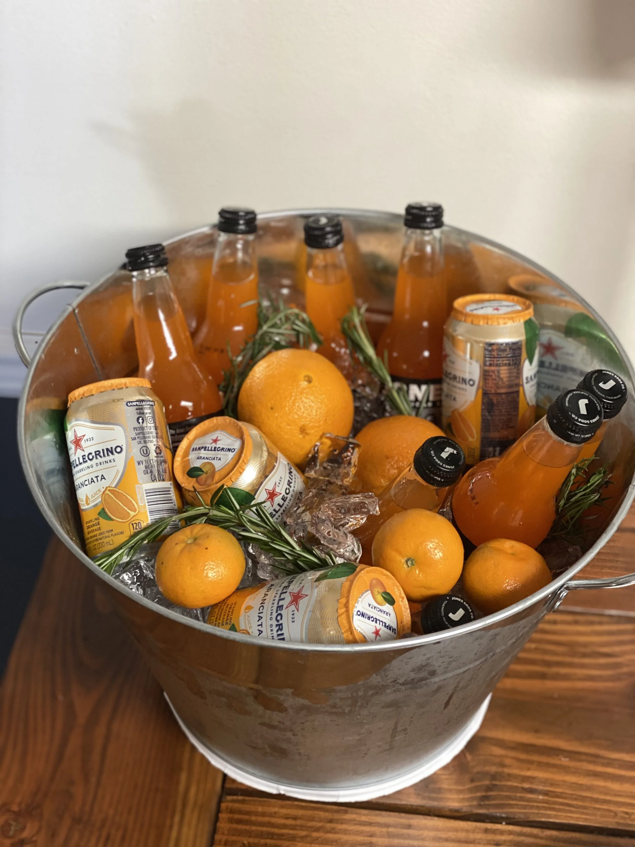 A metal bucket filled with ice, orange soda bottles, canned orange soda, fresh oranges, sprigs of rosemary, and ice cubes, placed on a wooden table.