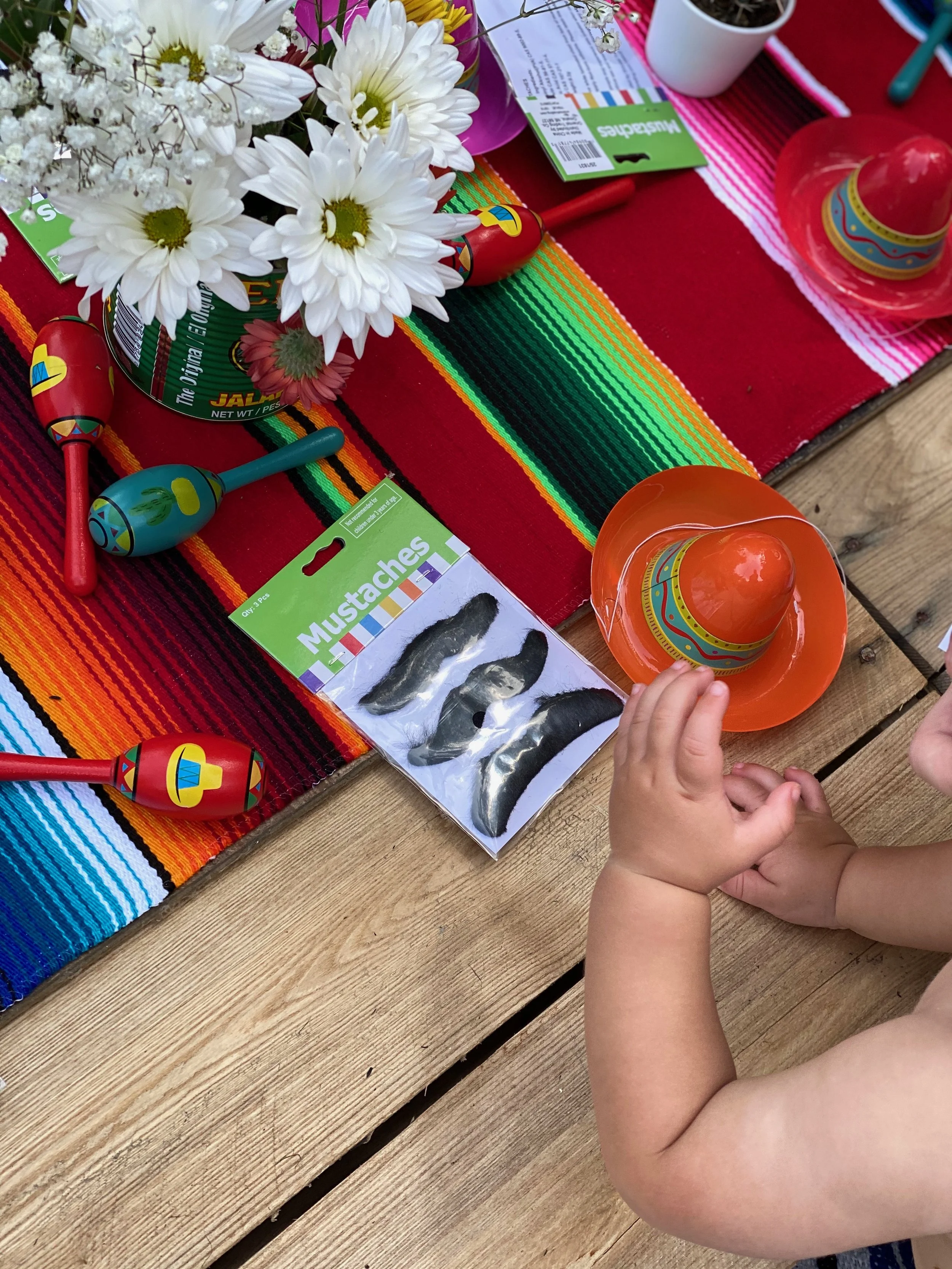 Child's hands reaching for a small orange and blue sombrero decor surrounded by colorful decorations, flowers, and a package of mustache stickers, placed on a vibrant multicolored striped tablecloth for a festive celebration.
