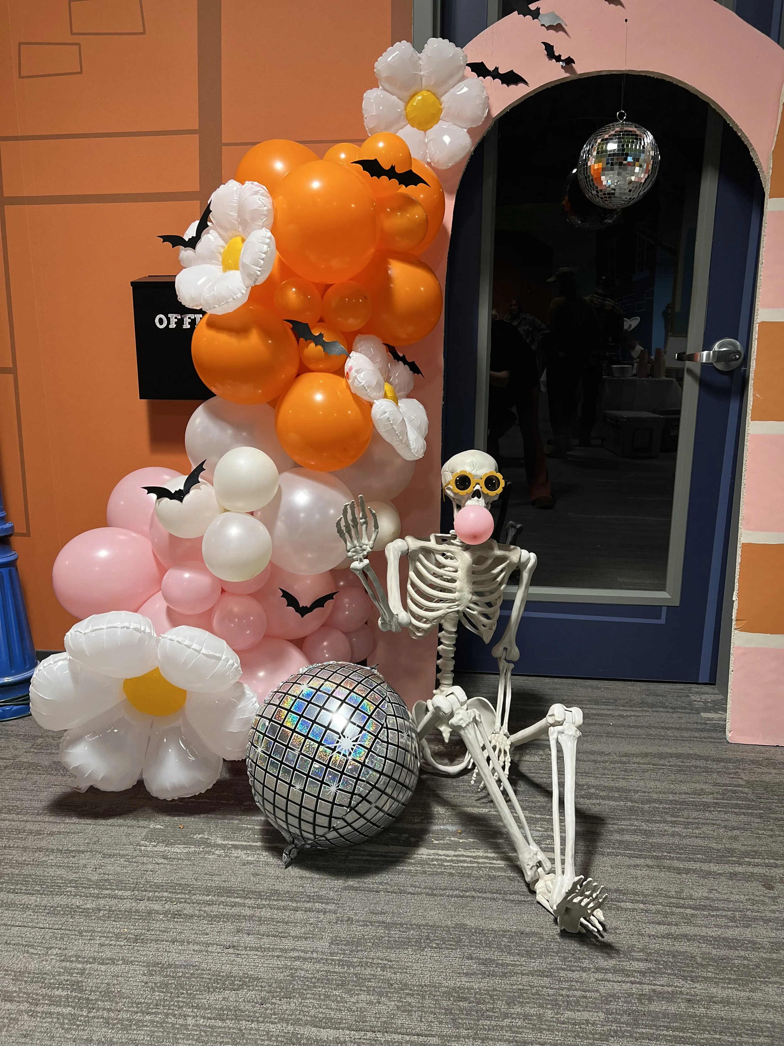 A Halloween decoration featuring a skeleton sitting on the floor with a pink clown nose and sunglasses, surrounded by orange, pink, and white balloons shaped like flowers and bats, with a silver disco ball hanging overhead.