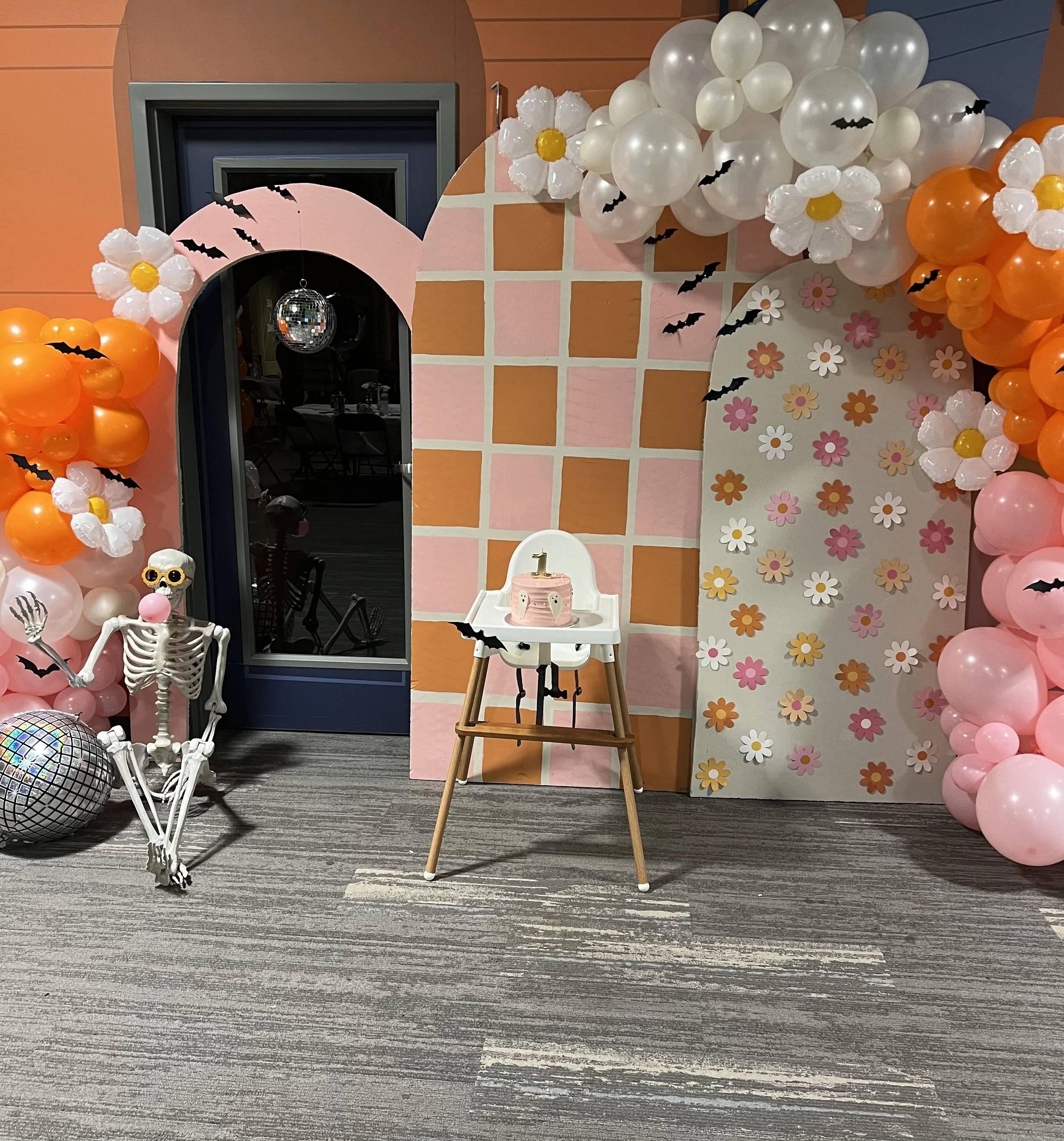 A decorated birthday party area with balloon arch, flowers, skeleton, disco ball, birthday cake with the number one candle, and Halloween-themed paper bats on the wall.