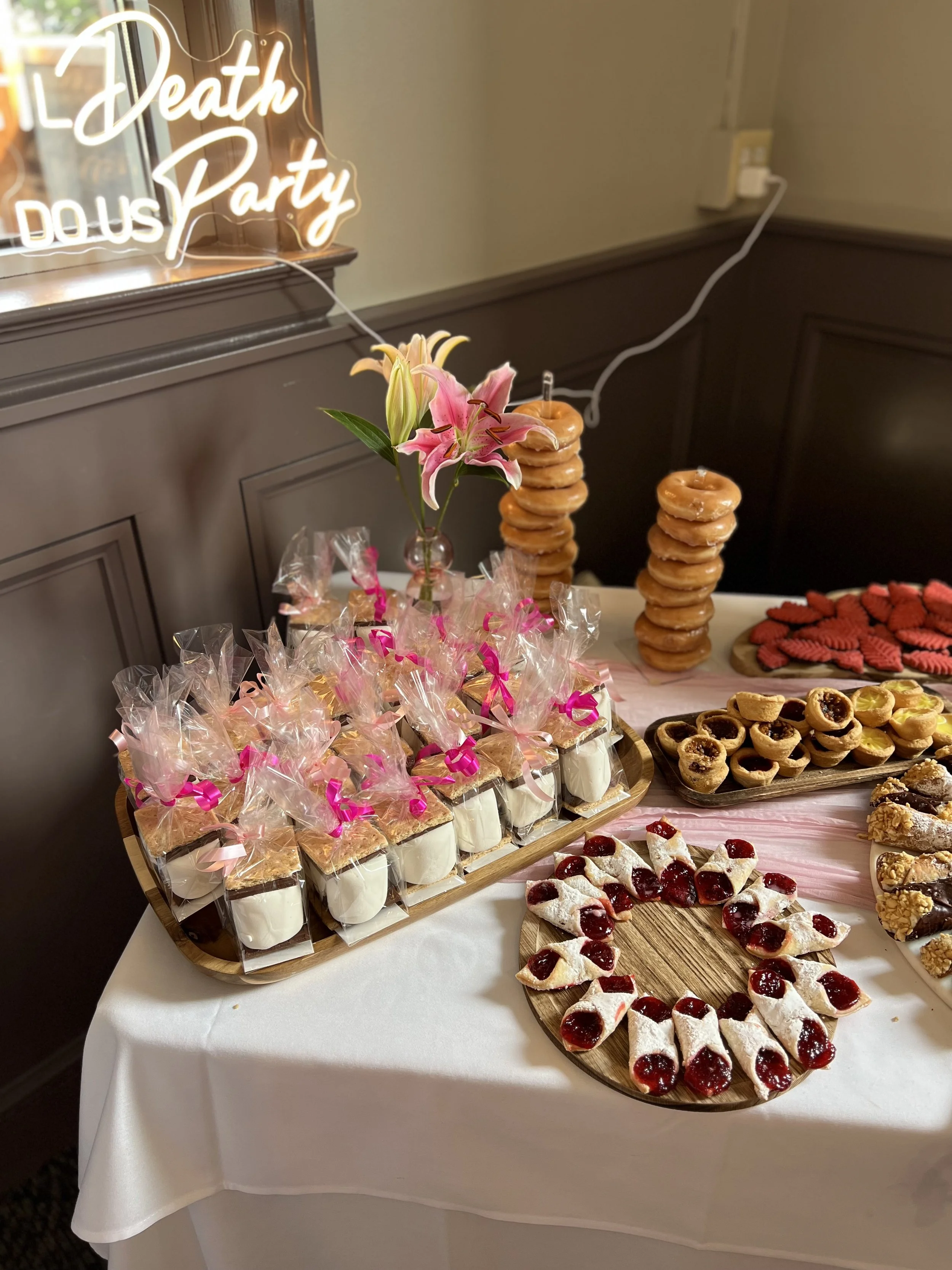 Dessert table with assorted pastries, cookies, and red fruit tarts. Pink lilies in a vase and a neon sign that says 'Death Doula Party' are on the table.