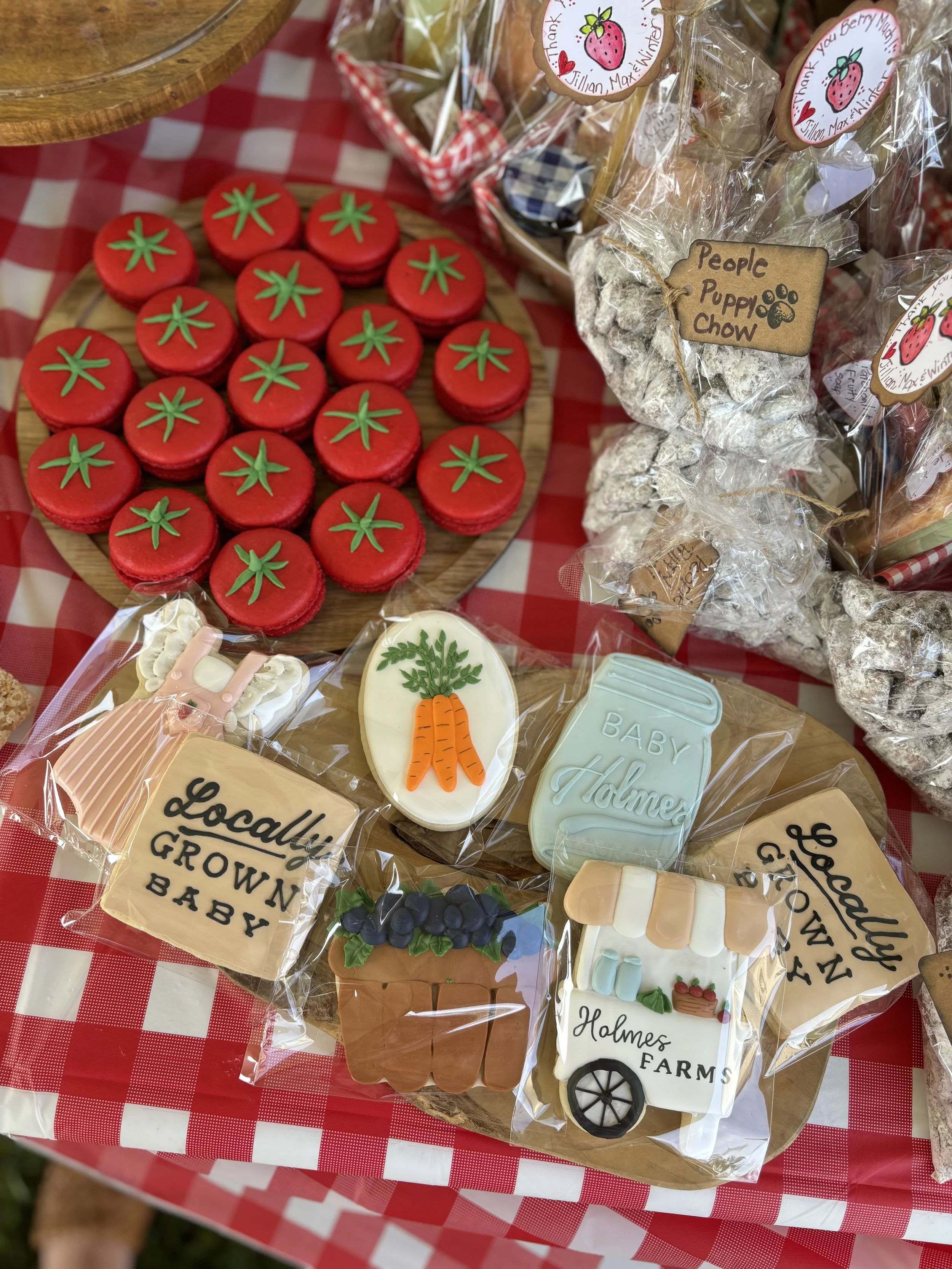 Assorted decorated cookies and baked goods on a table with a red and white checkered tablecloth. The cookies include red tomato-shaped ones, a carrot, a baby holly jar, a farm cart with holly, and signs saying "Locally Grown Baby" and "Holmes Farms."