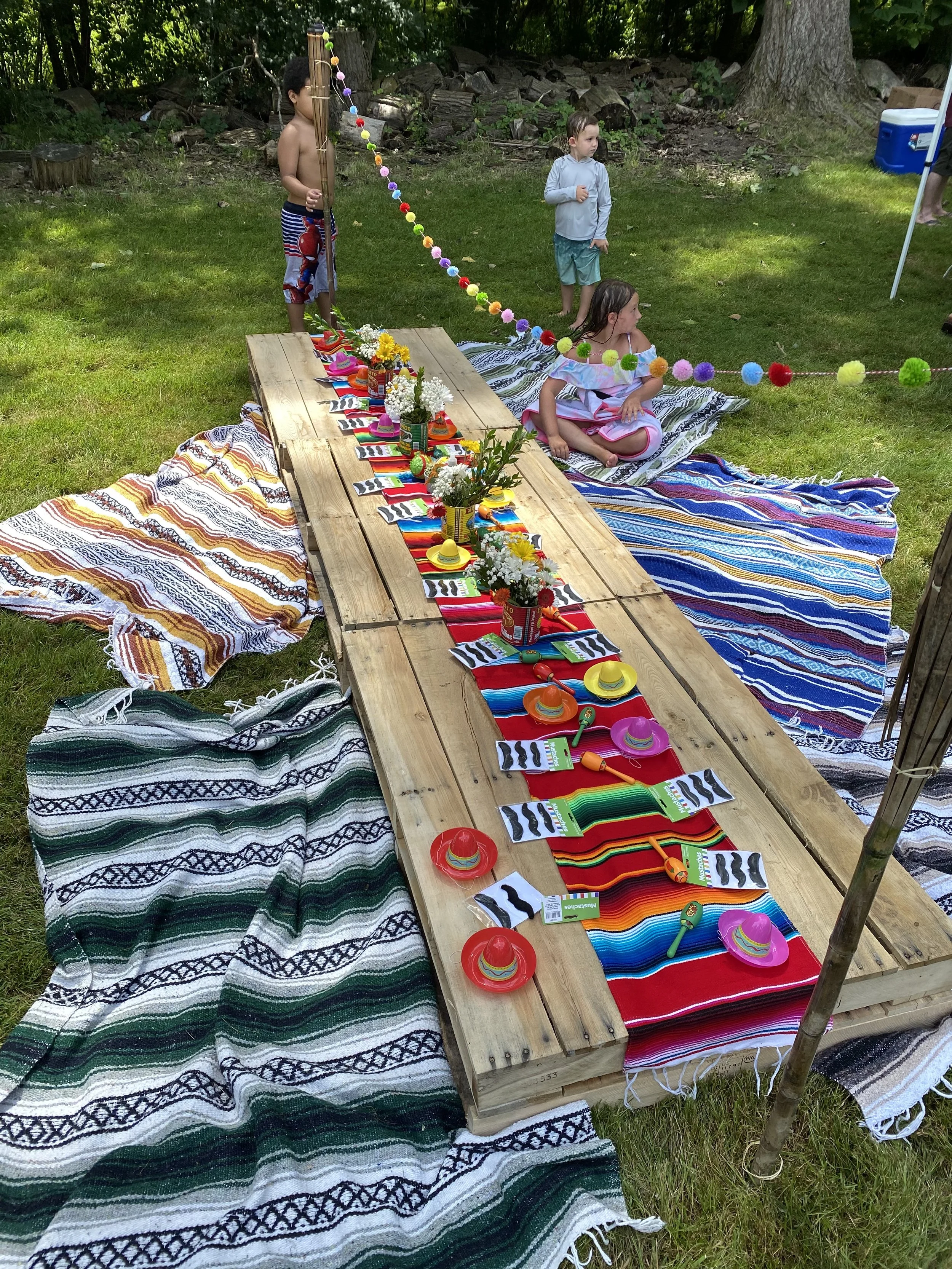 Colorful outdoor table setting with Mexican-themed decorations, sombreros, and striped table runners, surrounded by children at a backyard celebration.