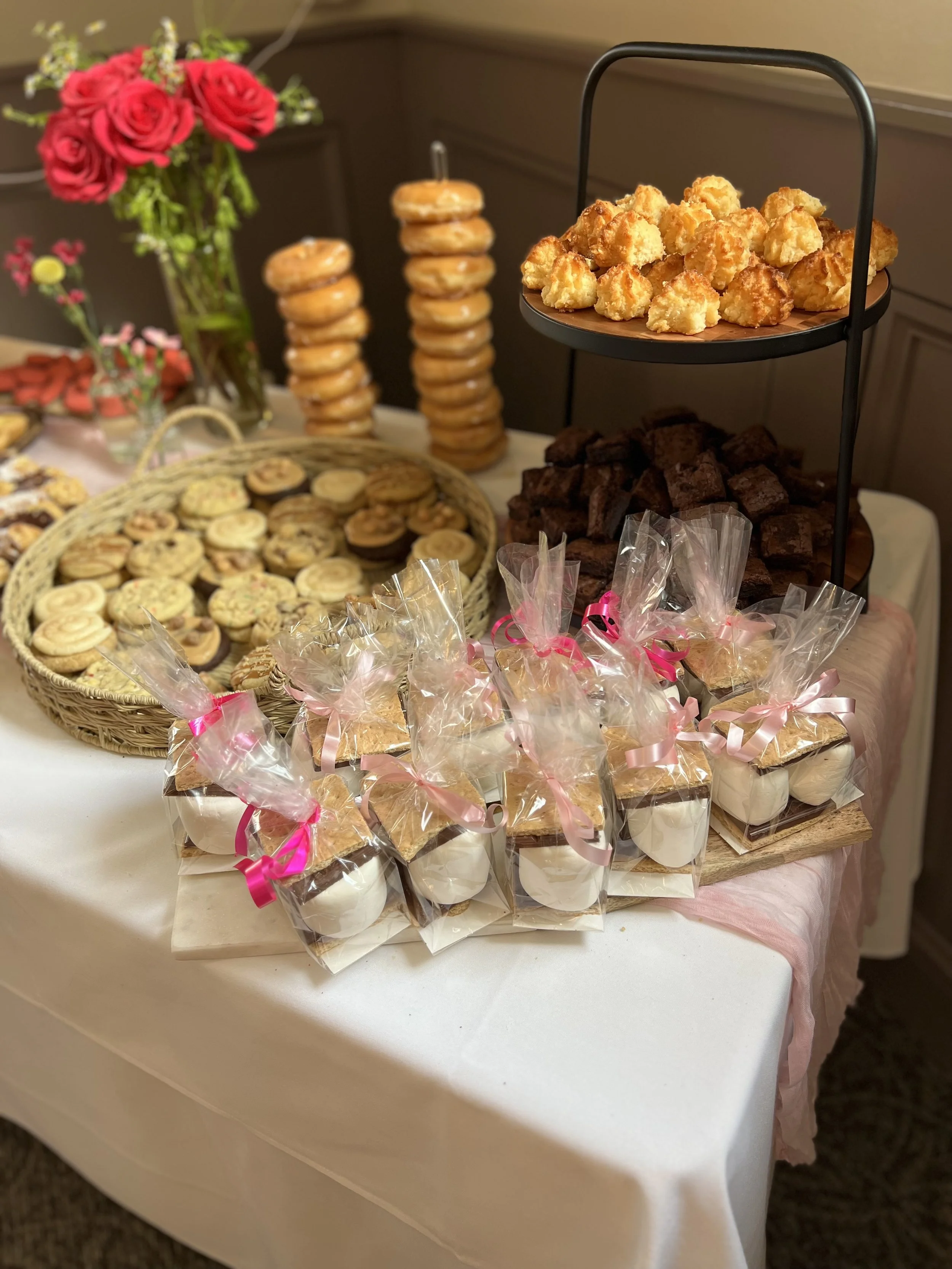 Arrangement of assorted baked goods including cookies, brownies, and mini parfaits, with a bouquet of red roses in the background.