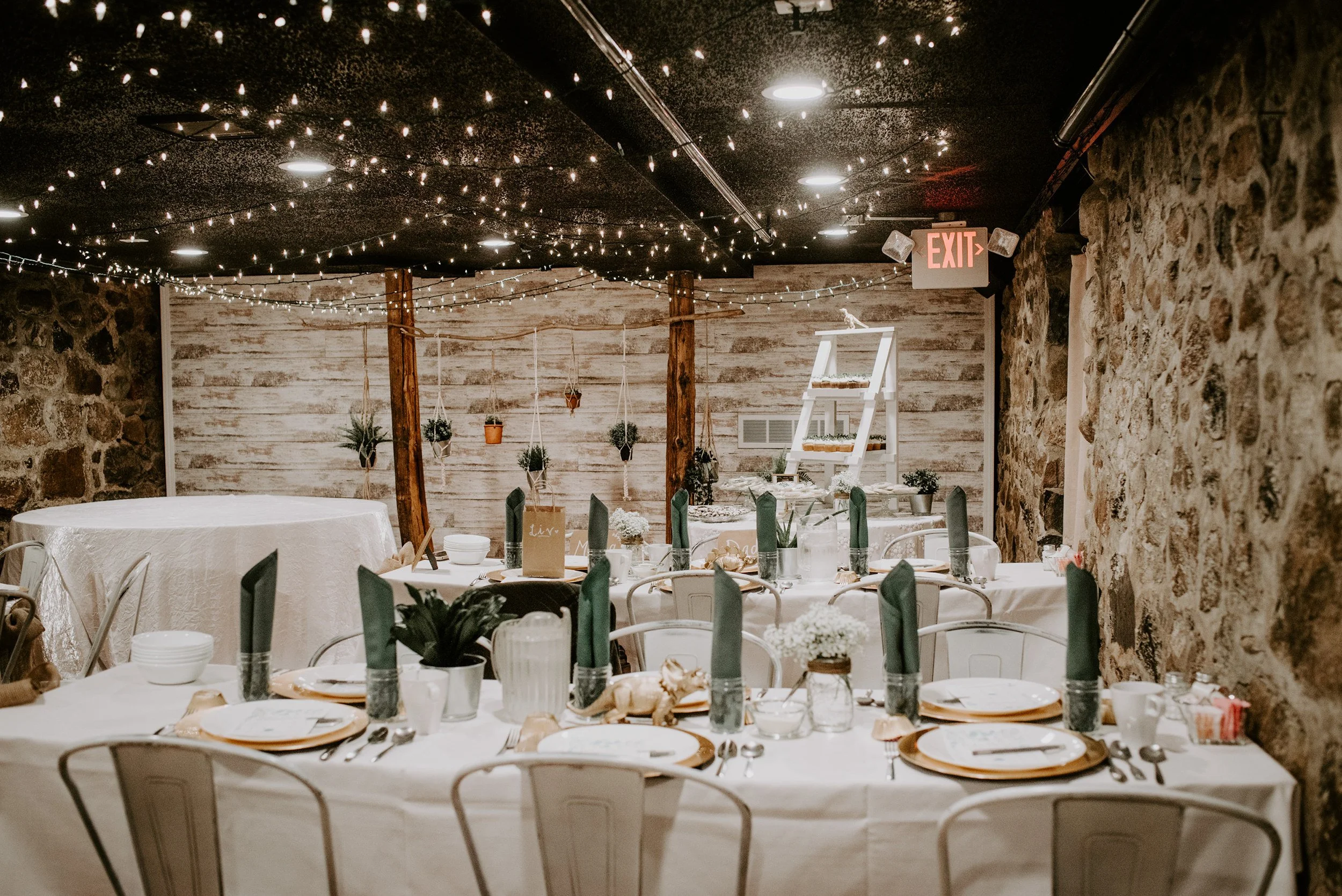 Decorated indoor dining area with string lights on ceiling, rustic stone and wood walls, round tables with white tablecloths, and place settings including plates, napkins, glasses, and utensils. Hanging potted plants and small floral arrangements are