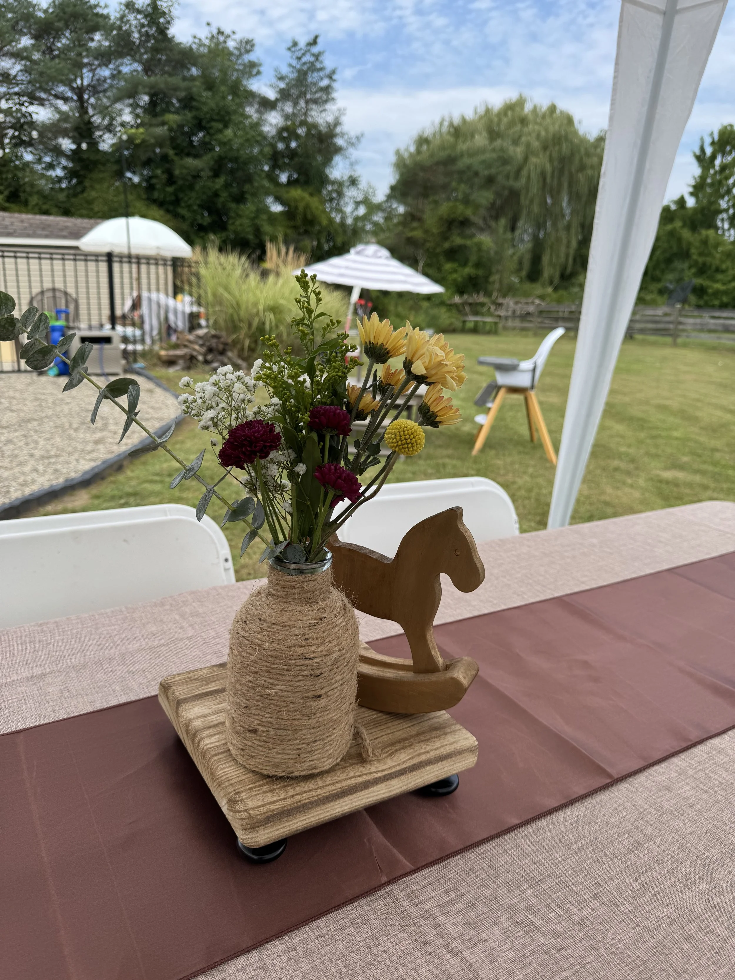 A floral arrangement in a wrapped vase on a wooden tray with a wooden horse decoration on a table. tablescape, hemp wrapped vases, outside baby shower, pony