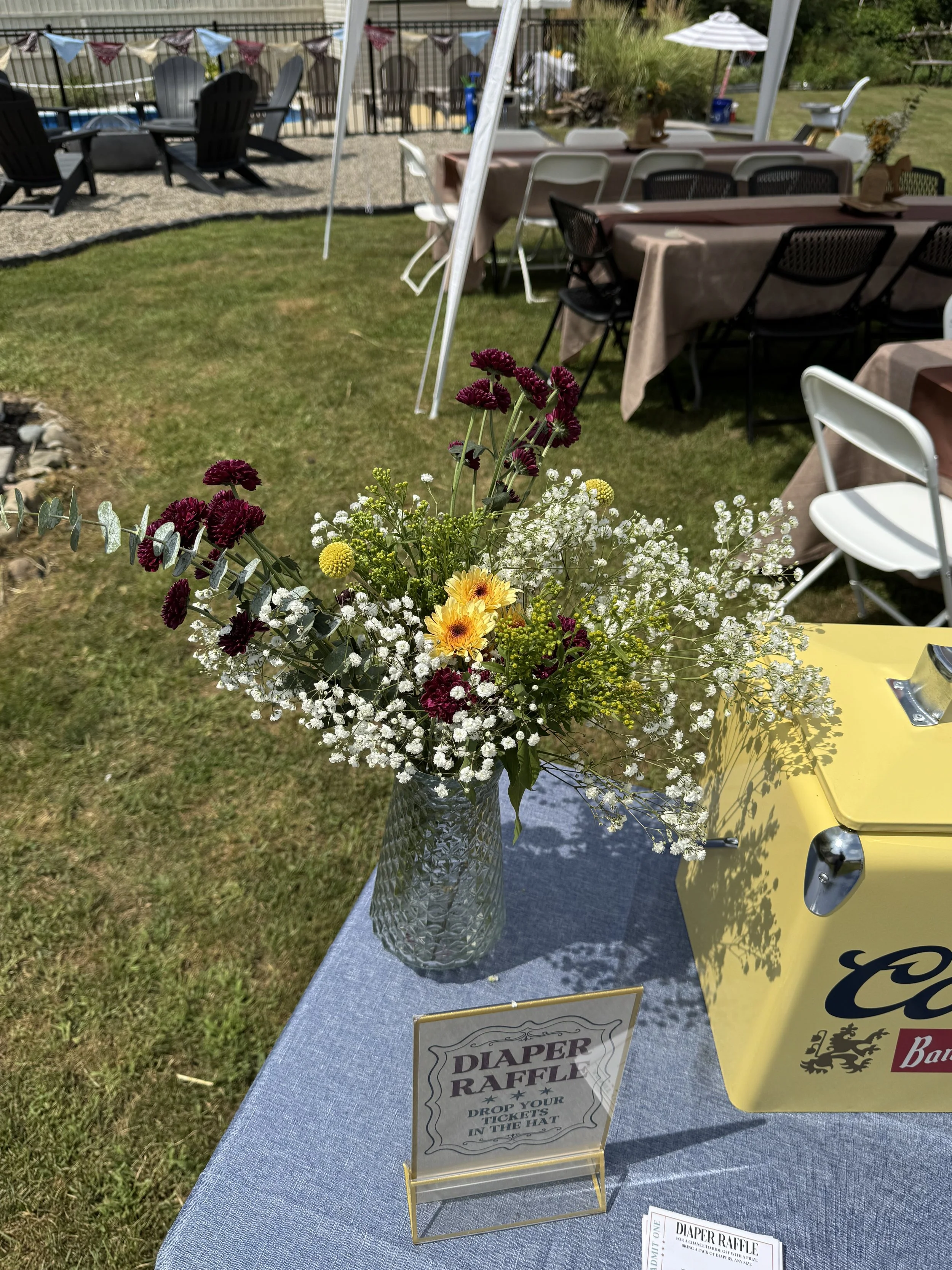 A vase of mixed flowers, including purple, yellow, and white blooms, is placed on a table at an outdoor event with other tables and chairs in the background. A sign on the table reads 'Diaper Raffle'.