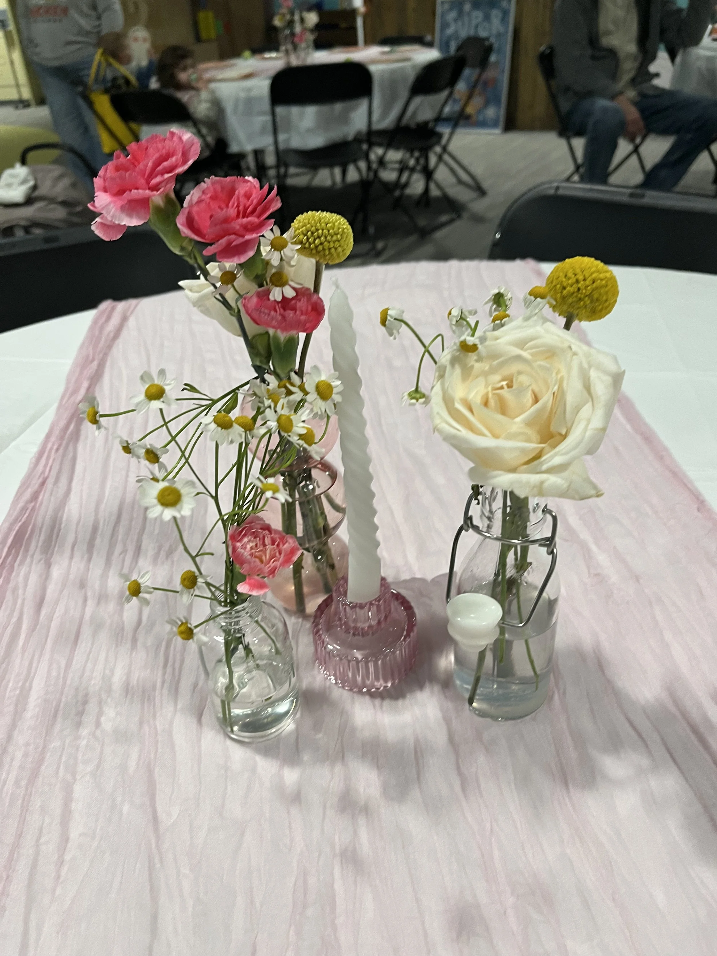 Table centerpiece with pink and white flowers, including pink carnations, daisies, and yellow Billy buttons, arranged in small glass vases with a pink candle in a pink holder and a white candle in a white holder, on a pink tablecloth at a gathering o