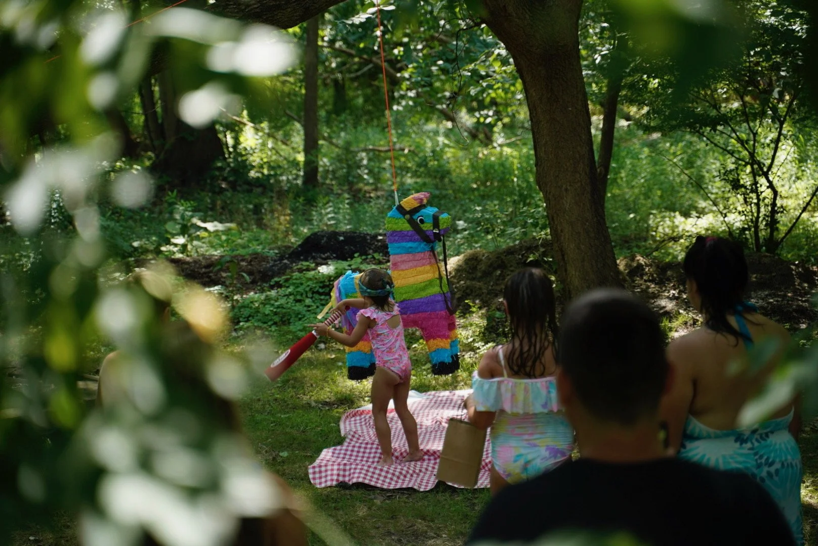 Children playing with a piñata shaped like a colorful llama in a lush green outdoor setting while adults watch.