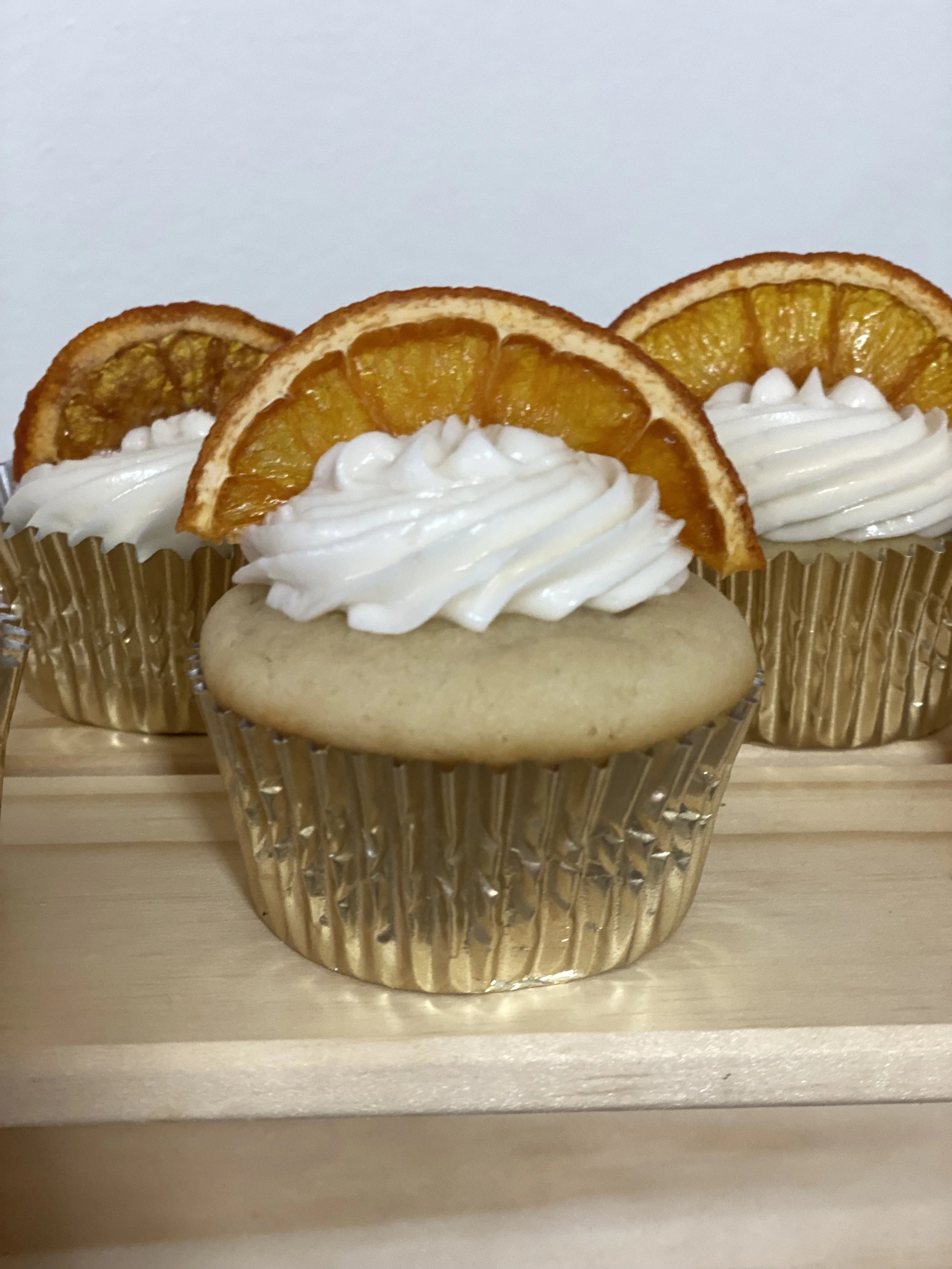 Cupcake topped with white frosting and an orange slice in a gold wrapper, displayed on a wooden surface.
