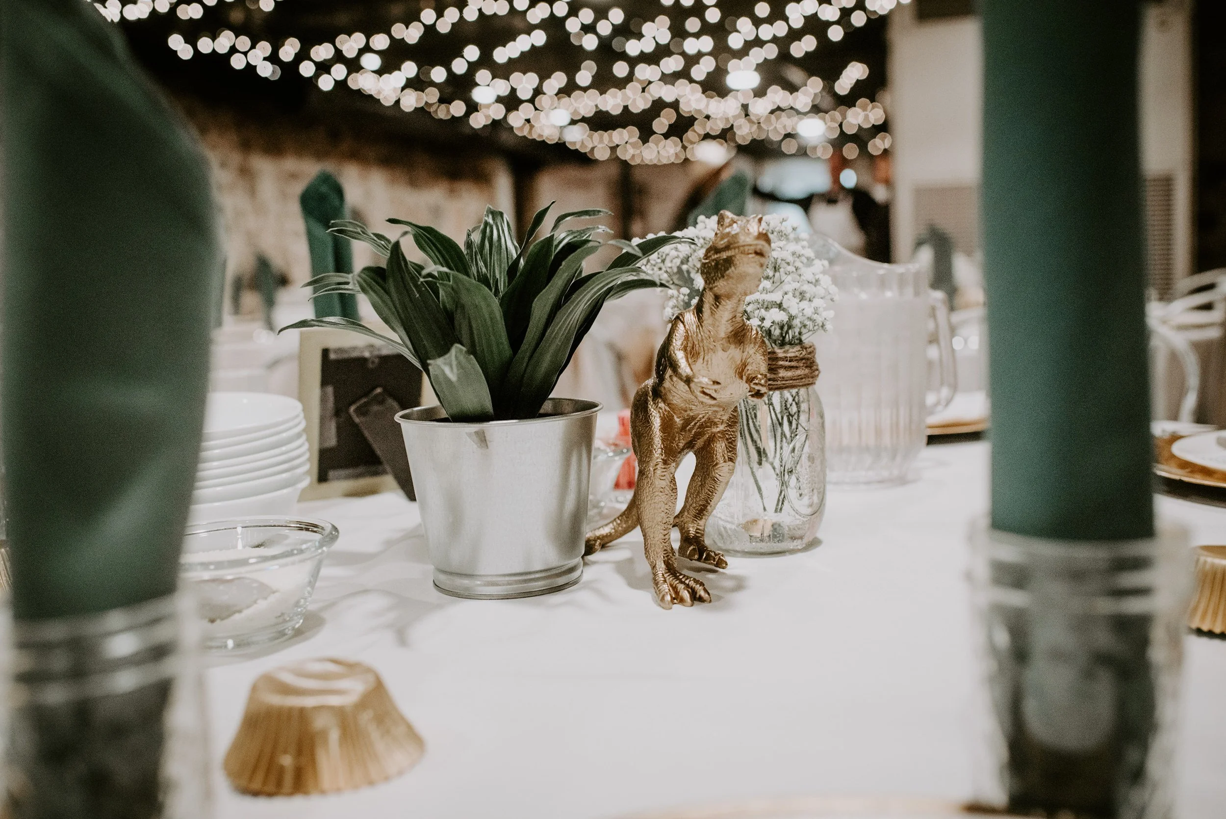Decorative table centerpiece with potted plants, a small dinosaur figurine, and flowers, surrounded by plates, glasses, and candles at a festive event.