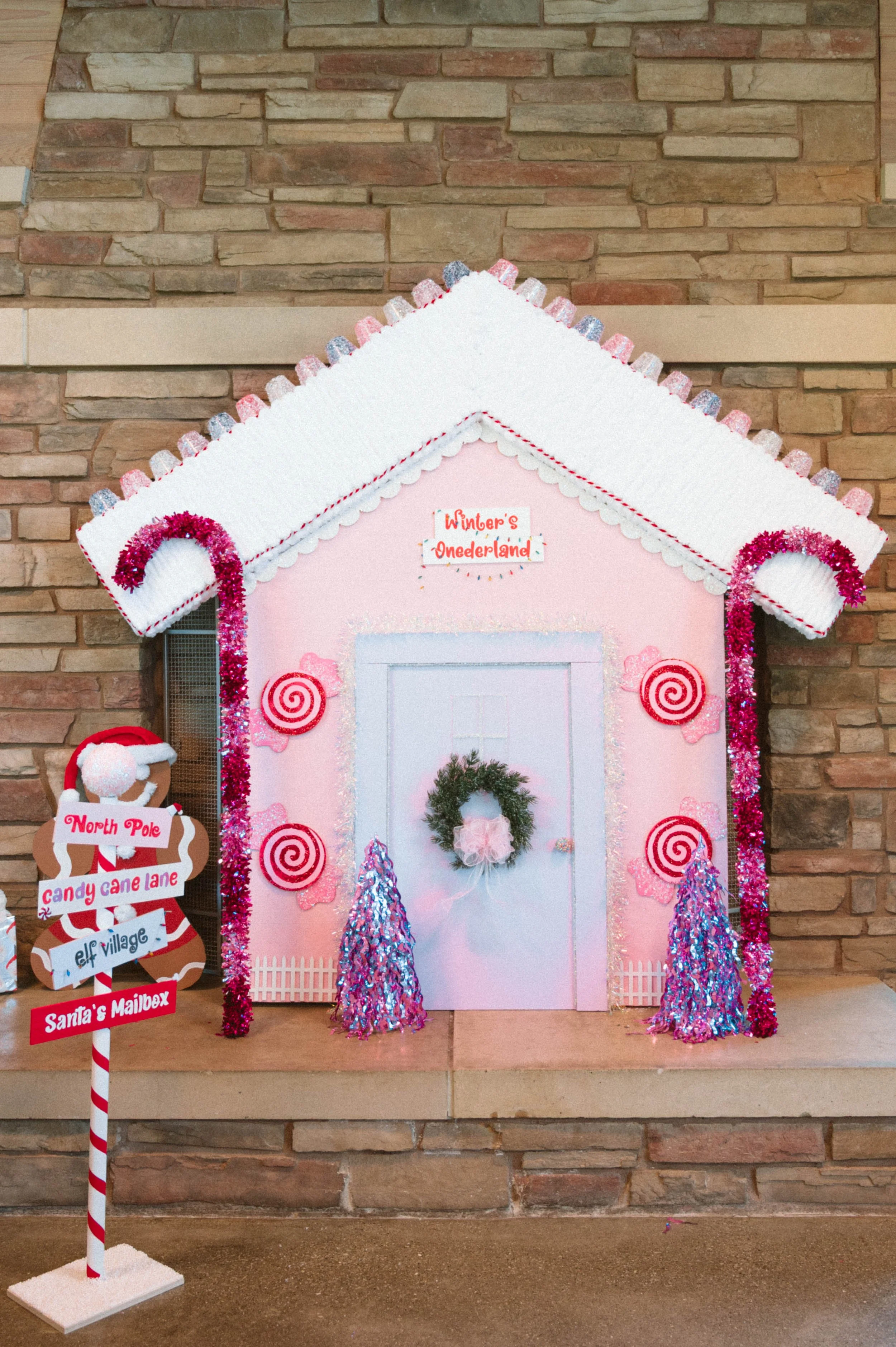 Decorated gingerbread house with pink walls, white roof, pink and purple tinsel, a green wreath on the door, and pink trees, set against a brick wall background, labeled "Winter's Wonderland." First birthday party theme, christmas theme