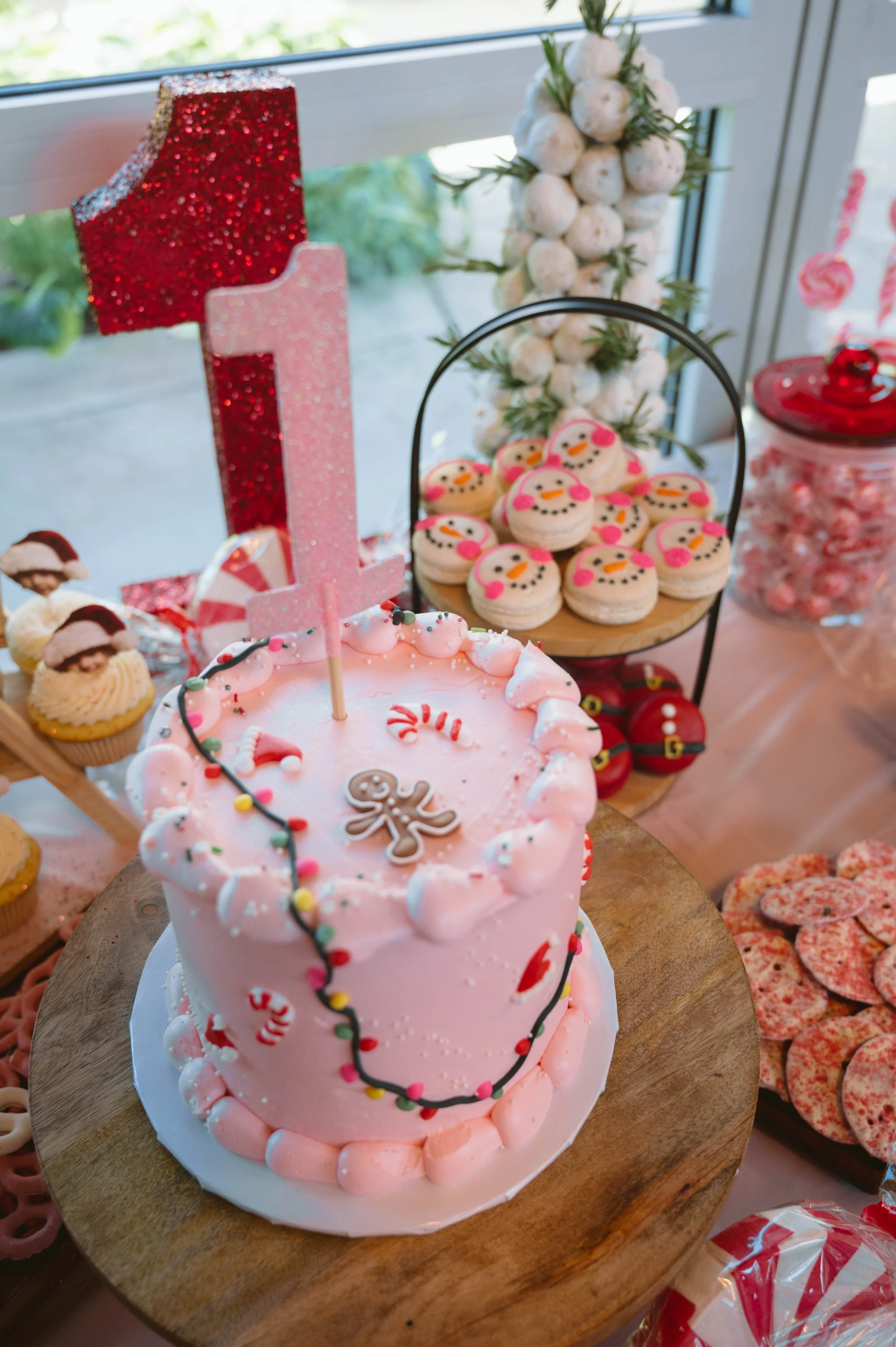 Pink birthday cake with icing decorations, topped with a number one candle, and surrounded by Christmas-themed cookies and cupcakes on a table.