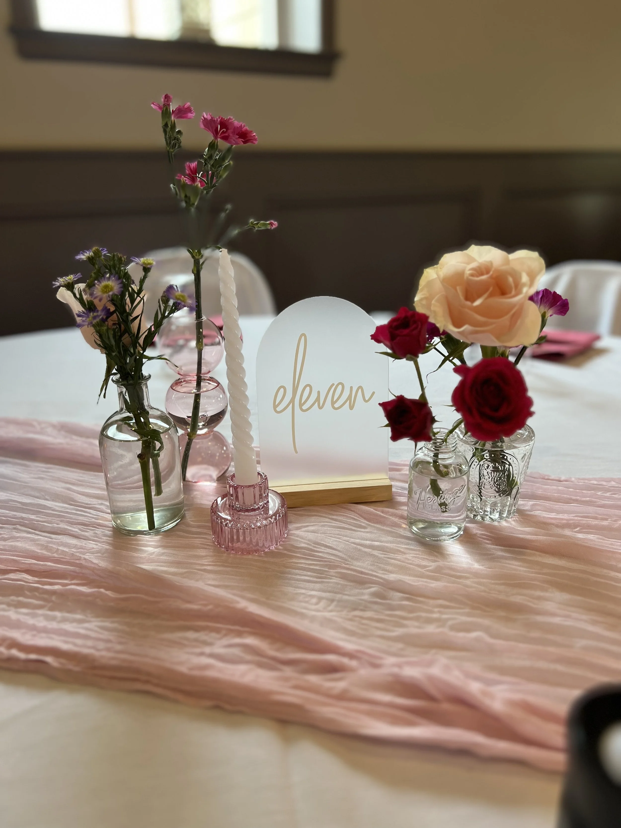 Table centerpiece with small glass vases holding colorful flowers, a white decorative number 'eleven' sign, a pink candle holder with a twisted white candle, and a pink fabric table runner.