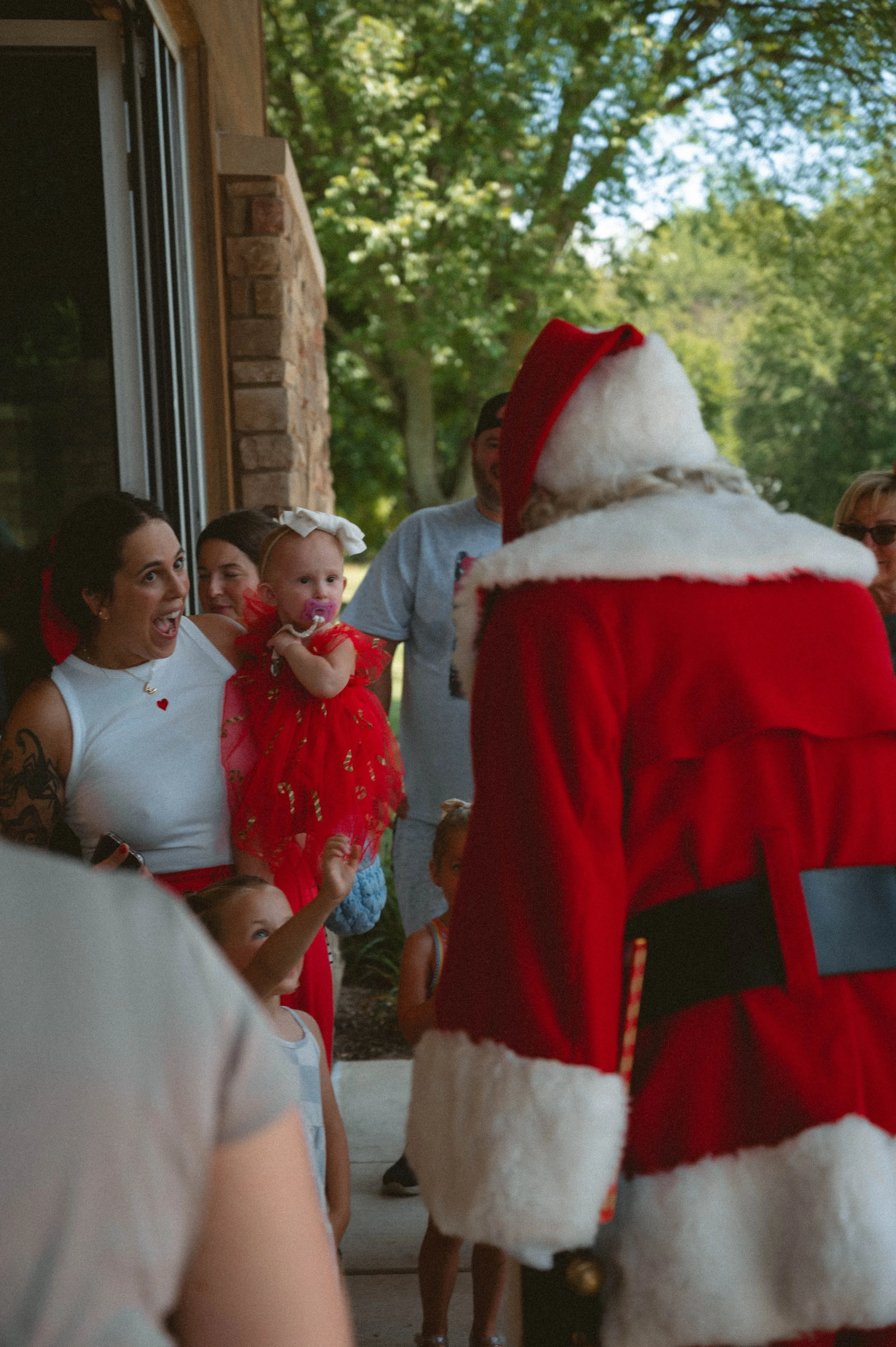 Children and adults excitedly greeting Santa Claus. The children are smiling; one girl in a red dress and a large white bow on her head is being held up, while others watch and wave. Santa, dressed in traditional red and white, is facing the group wi