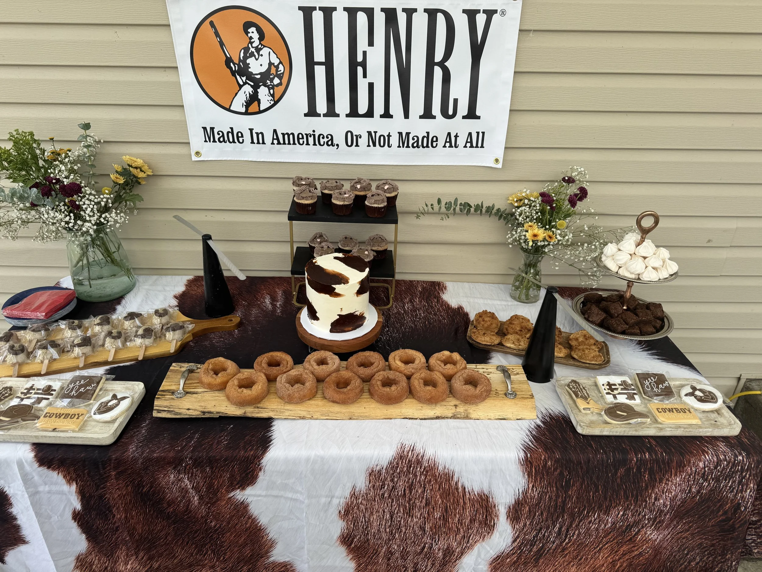 A table with a cowhide tablecloth displaying assorted baked goods, including donuts, cupcakes, cookies, and decorated cookies. The background features a banner that reads "Henry Made In America, Or Not Made At All," along with floral arrangements in