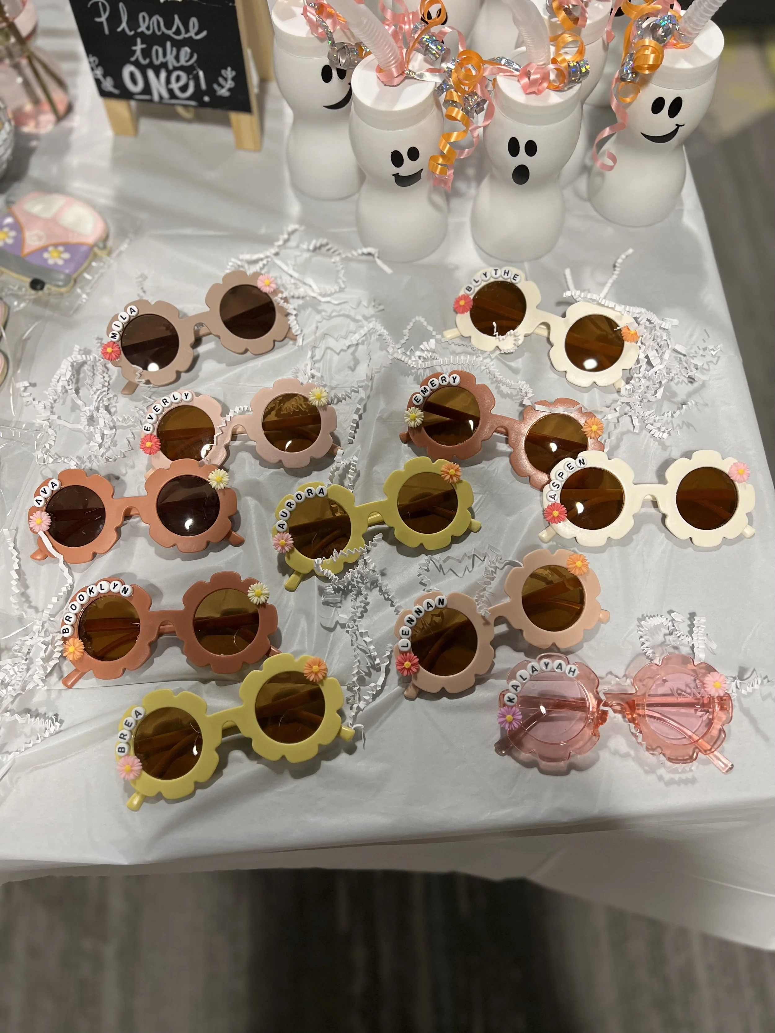 Colorful flower-shaped sunglasses with pageant-style name tags on the lenses, arranged on a table with shredded paper and white ghost-shaped bottles with curly ribbon decorations, at a celebration event.