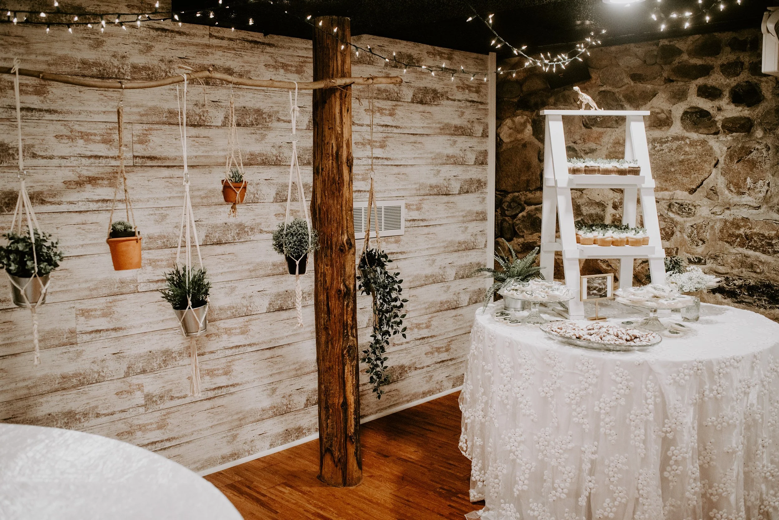 A rustic indoor setting with a wooden wall, string lights, potted plants hanging on a rope, a wooden post, and a white tiered display table with floral arrangements and small framed pictures, on a round table with a lace tablecloth.