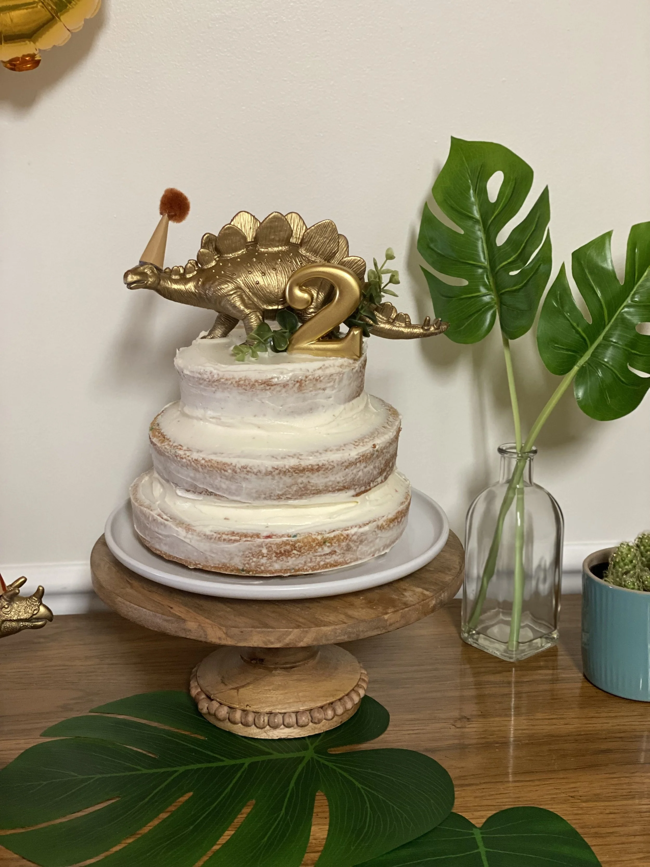 A birthday cake with a dinosaur and a gold number 2 topper, decorated with greenery, on a wooden cake stand, with large green fern leaves and smaller plants nearby.