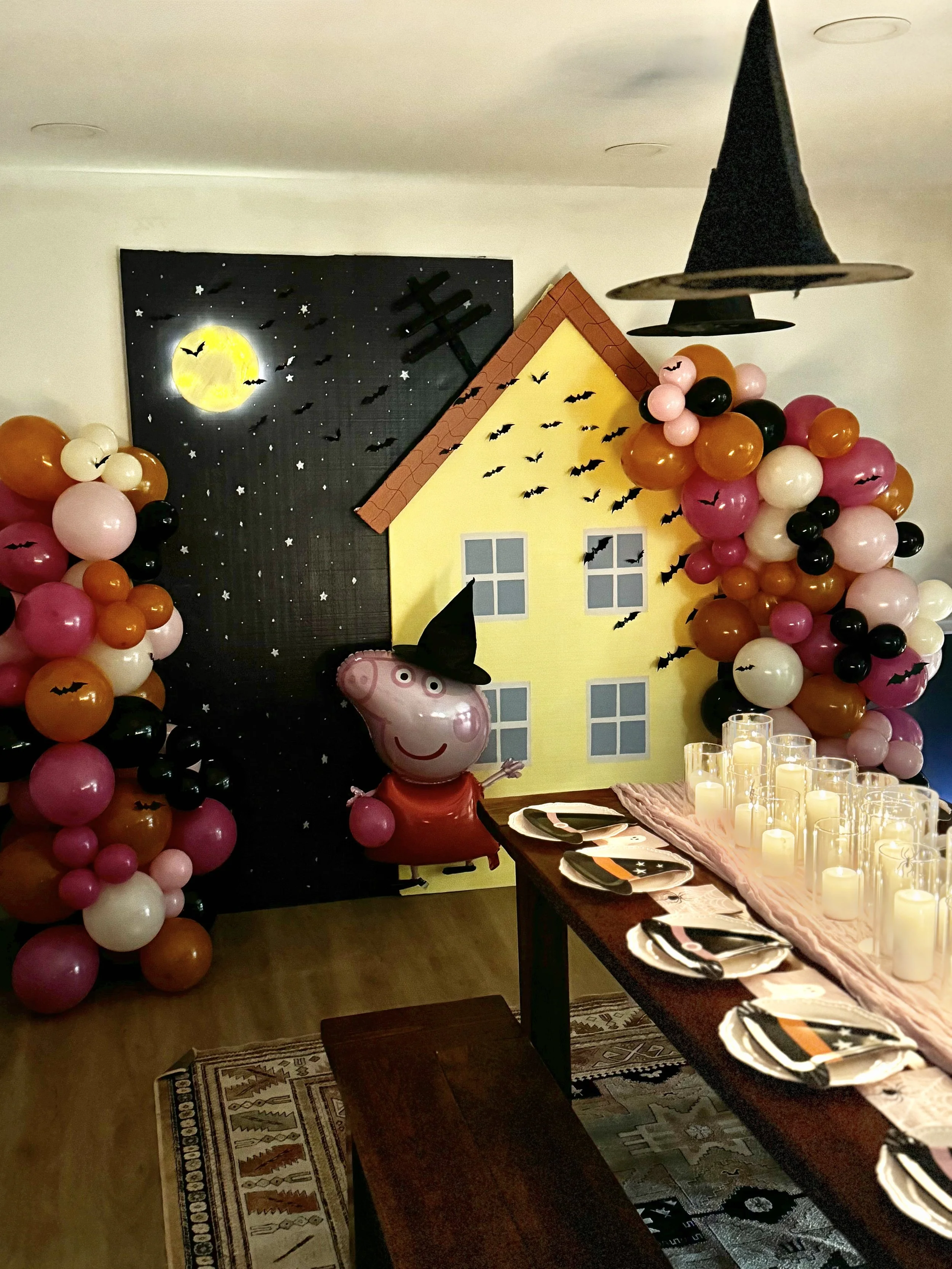 A Halloween-themed party room with a Peppa Pig balloon dressed as a witch, colorful balloon decorations with bats, a house backdrop, a moon and stars on a black sky, a witch hat hanging from the ceiling, and a table set with candles, plates, and napk