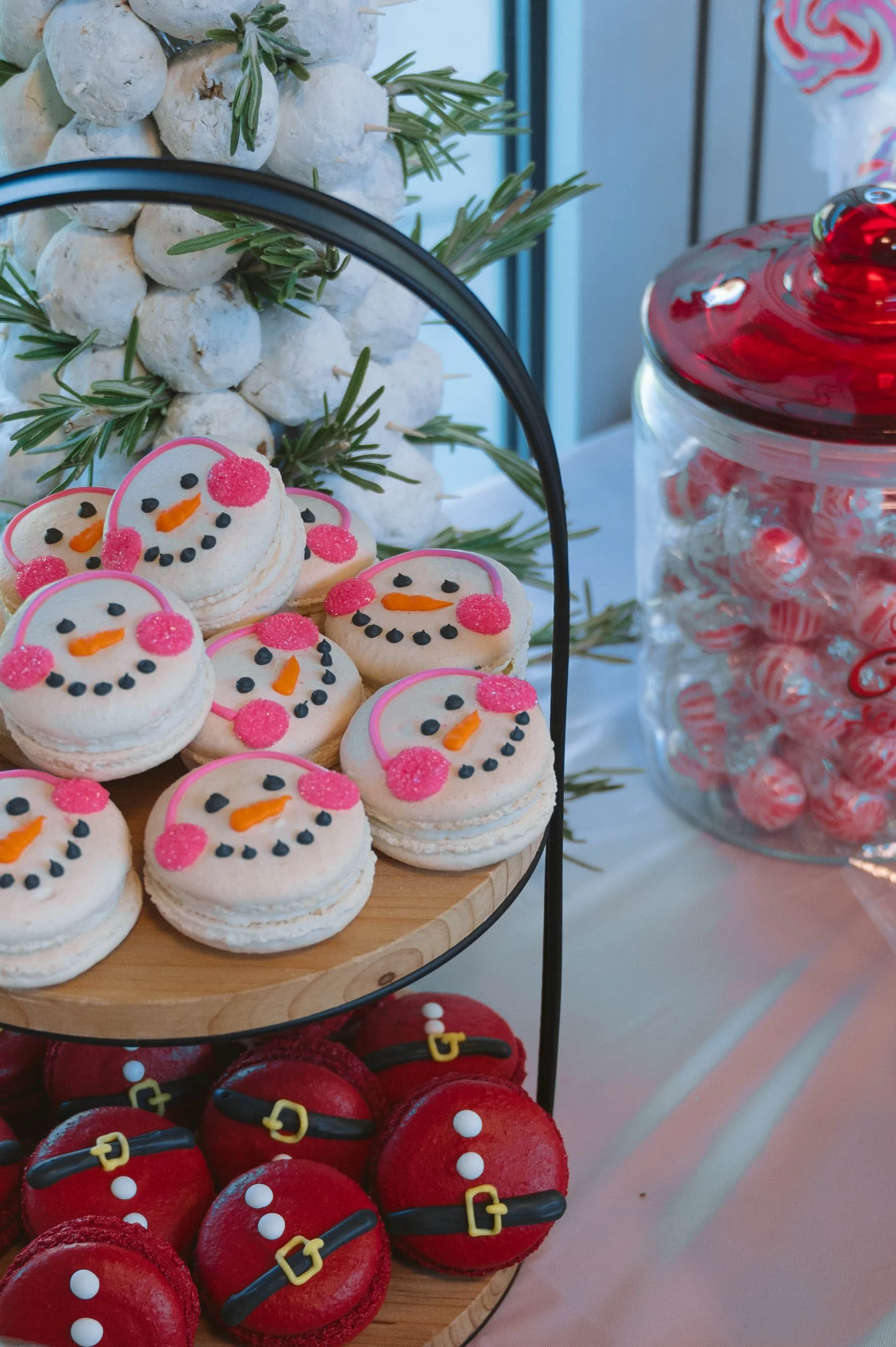 Snowman decorated cookies with pink cheeks, orange noses, and black eyes, alongside red Christmas cookies with belt decorations, displayed on a two-tiered stand near a jar of peppermint candies.